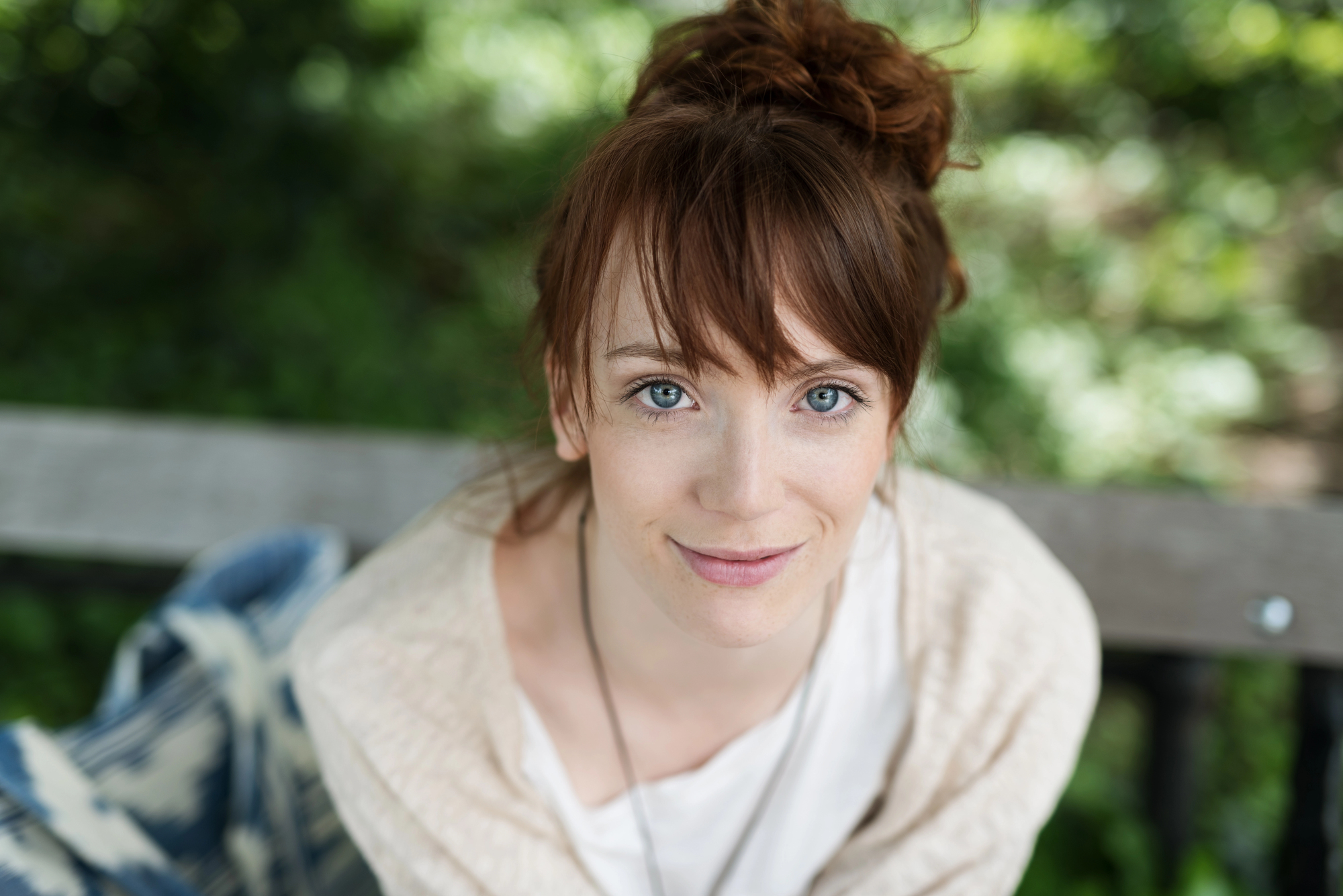 A woman with red hair and blue eyes is sitting outdoors on a wooden bench. She has her hair tied up in a messy bun and is wearing a light-colored top and a necklace. The background is blurred with greenery. She looks into the camera with a slight smile.