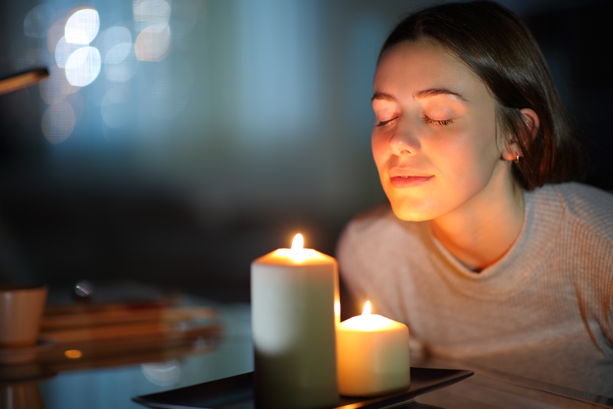 A young woman with eyes closed smiles softly while leaning close to two large lit candles on a dining table. The background is blurred, featuring a cozy indoor setting with warm ambient lighting.