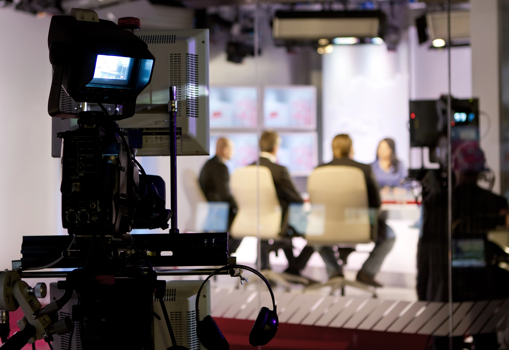 A TV camera focuses on a brightly lit studio set where four people are sitting around a circular table discussing. The camera and its equipment are in the foreground, partially obscuring the view of the people. Monitors and screens are visible in the background.