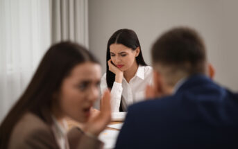 A woman looks troubled and sad while sitting at a table. In the foreground, two people are whispering to each other. The scene suggests the woman may be feeling left out or gossiped about by the others. The setting appears to be an office or meeting room.