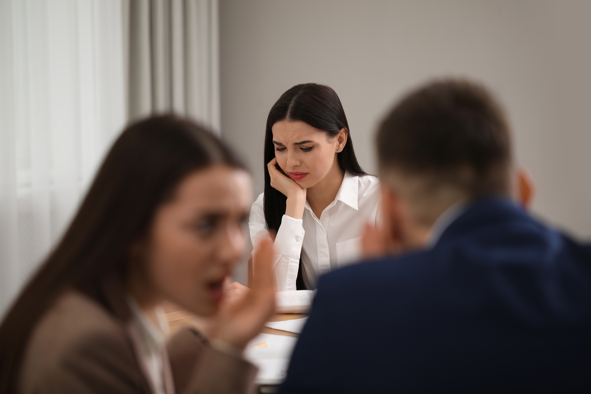 A woman looks troubled and sad while sitting at a table. In the foreground, two people are whispering to each other. The scene suggests the woman may be feeling left out or gossiped about by the others. The setting appears to be an office or meeting room.