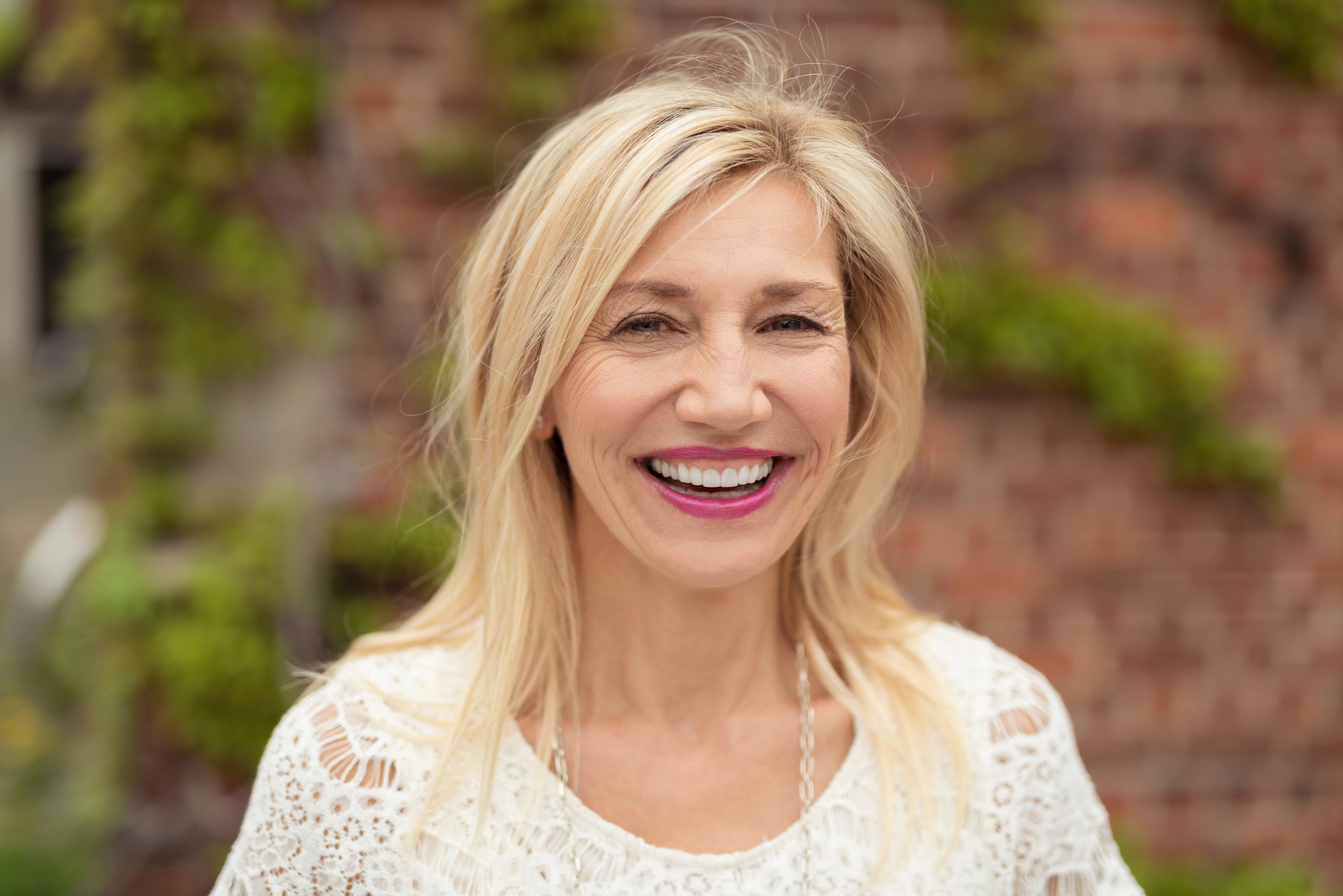 A smiling woman with long blonde hair is standing outdoors in front of a brick wall with green vines. She is wearing a white lace top and has a joyful expression.