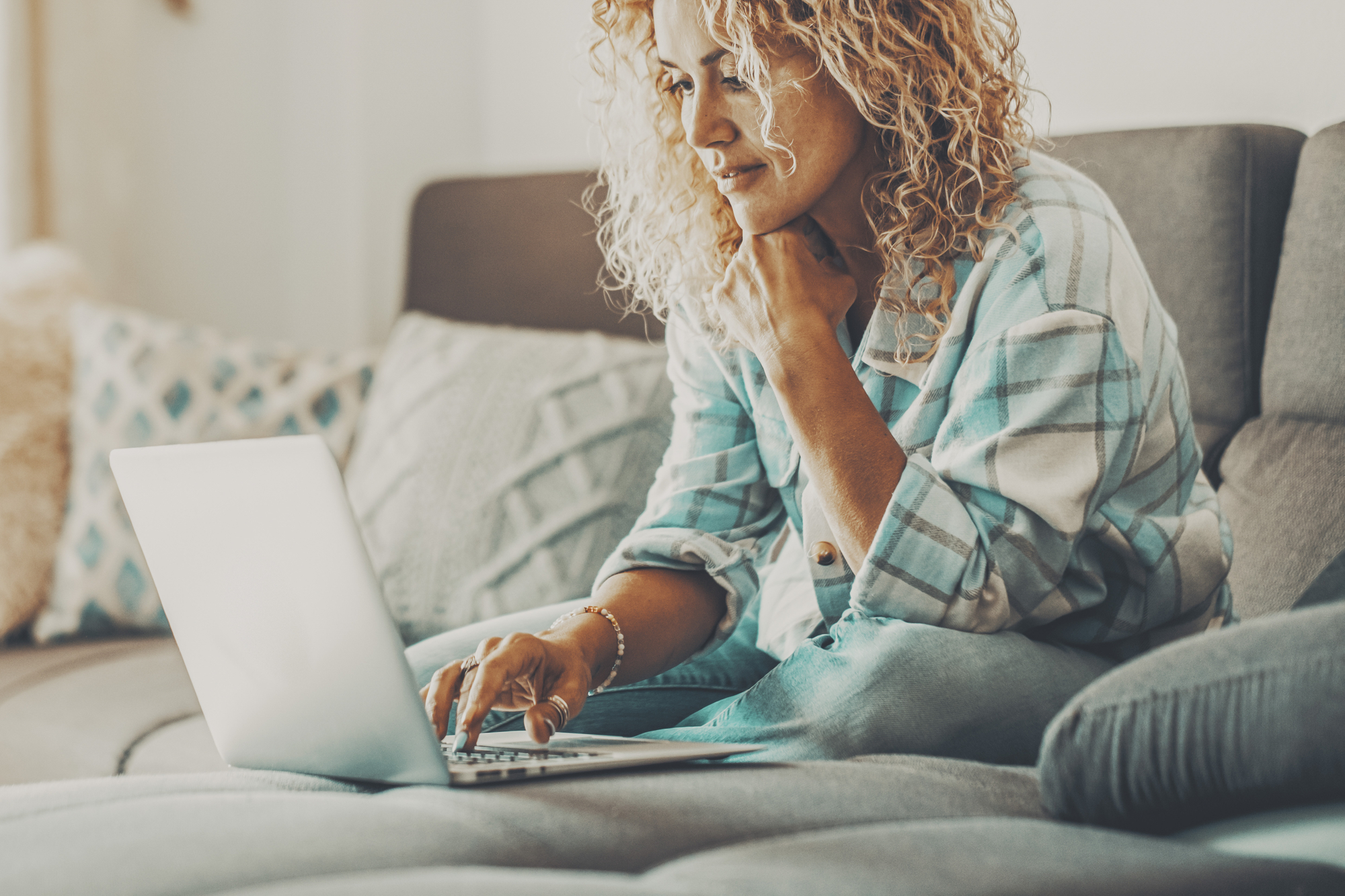 A person with curly hair sits on a couch while typing on a laptop. They are wearing a blue and white plaid shirt and seem focused on the screen. The setting appears to be a cozy living room with cushions and pillows in the background.