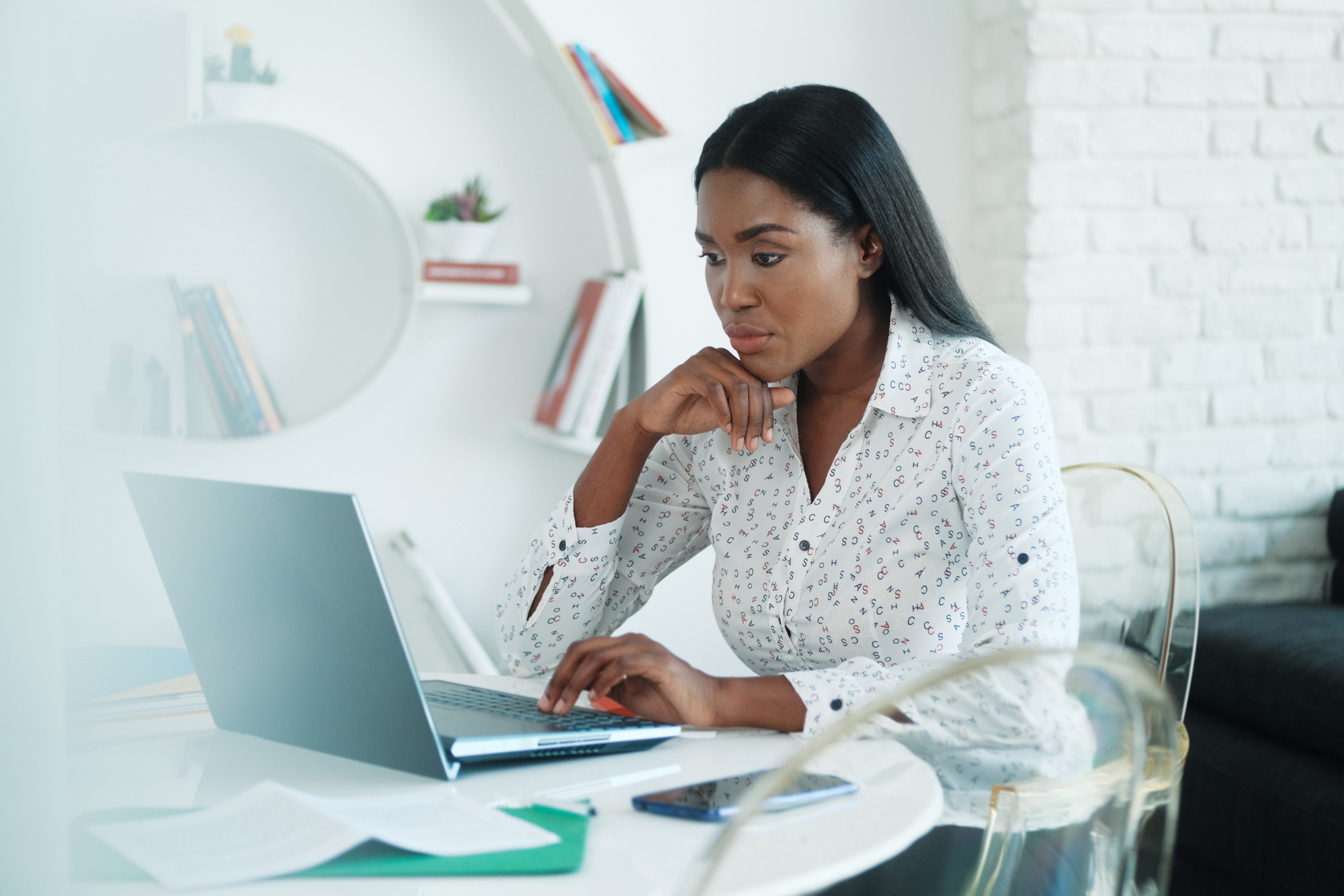 A woman sits at a round white table, intently working on her laptop. She wears a white patterned blouse and rests her chin on her hand. Papers, a pen, and a smartphone lie on the table. In the background is a white wall with circular shelves holding books and plants.