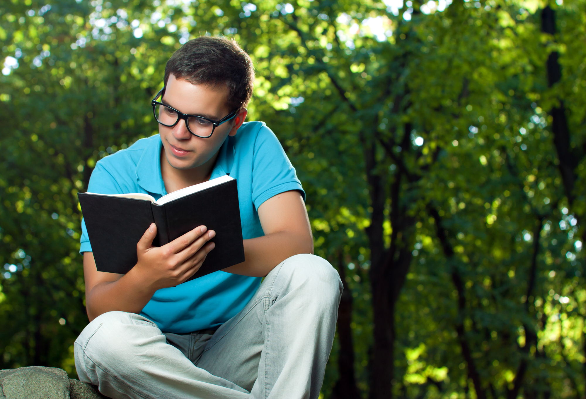 Young man with short dark hair and glasses sits outdoors reading a black book. He wears a blue polo shirt and light-colored pants. The background is lush with green trees, suggesting a peaceful, wooded setting. The lighting is bright and natural.