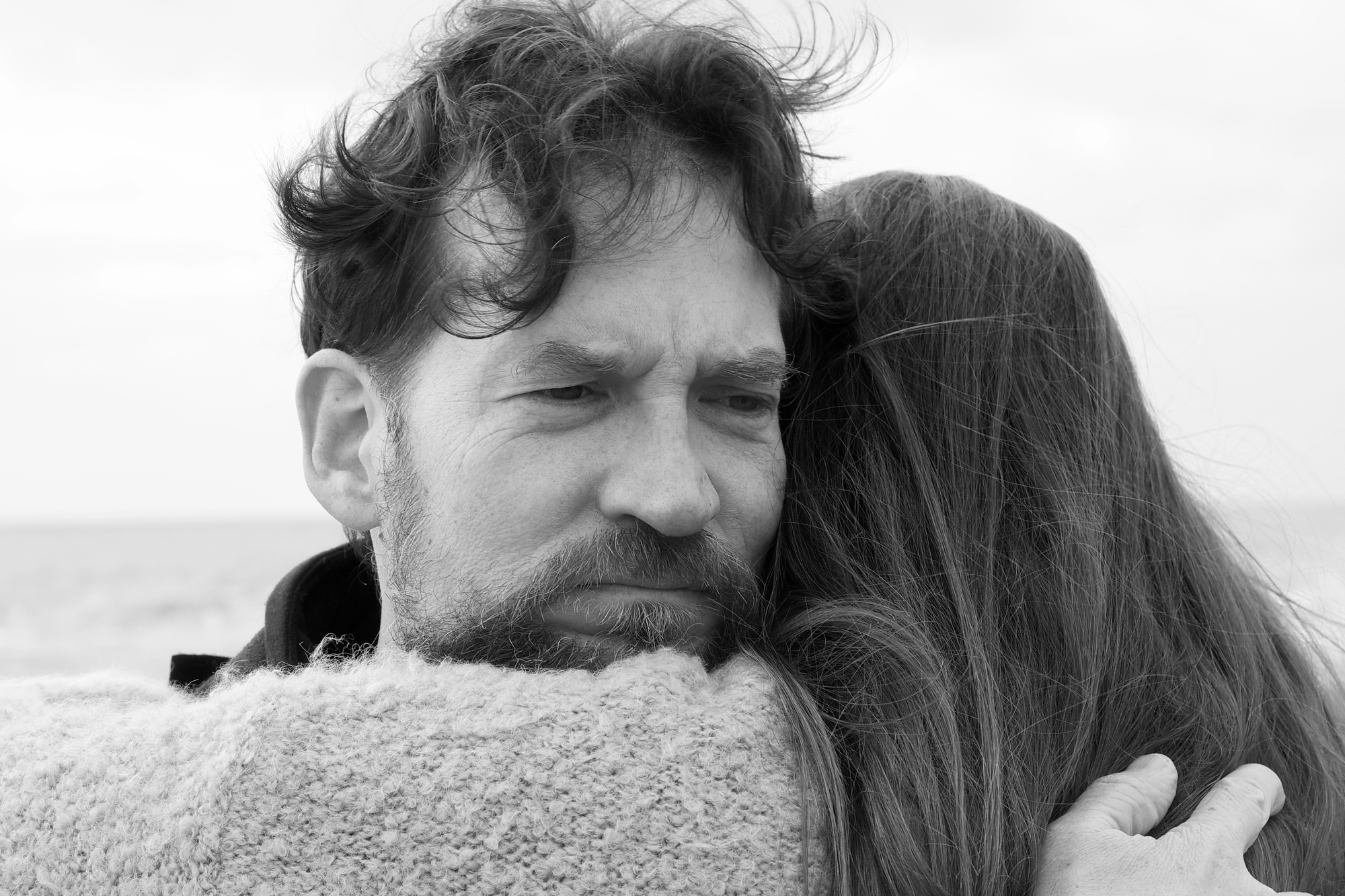 A black and white photo shows a man with tousled hair and a beard, embracing a woman from behind. The man's expression appears contemplative, while the woman, facing away, rests her head on his shoulder. The background is a blurred, serene outdoor setting.