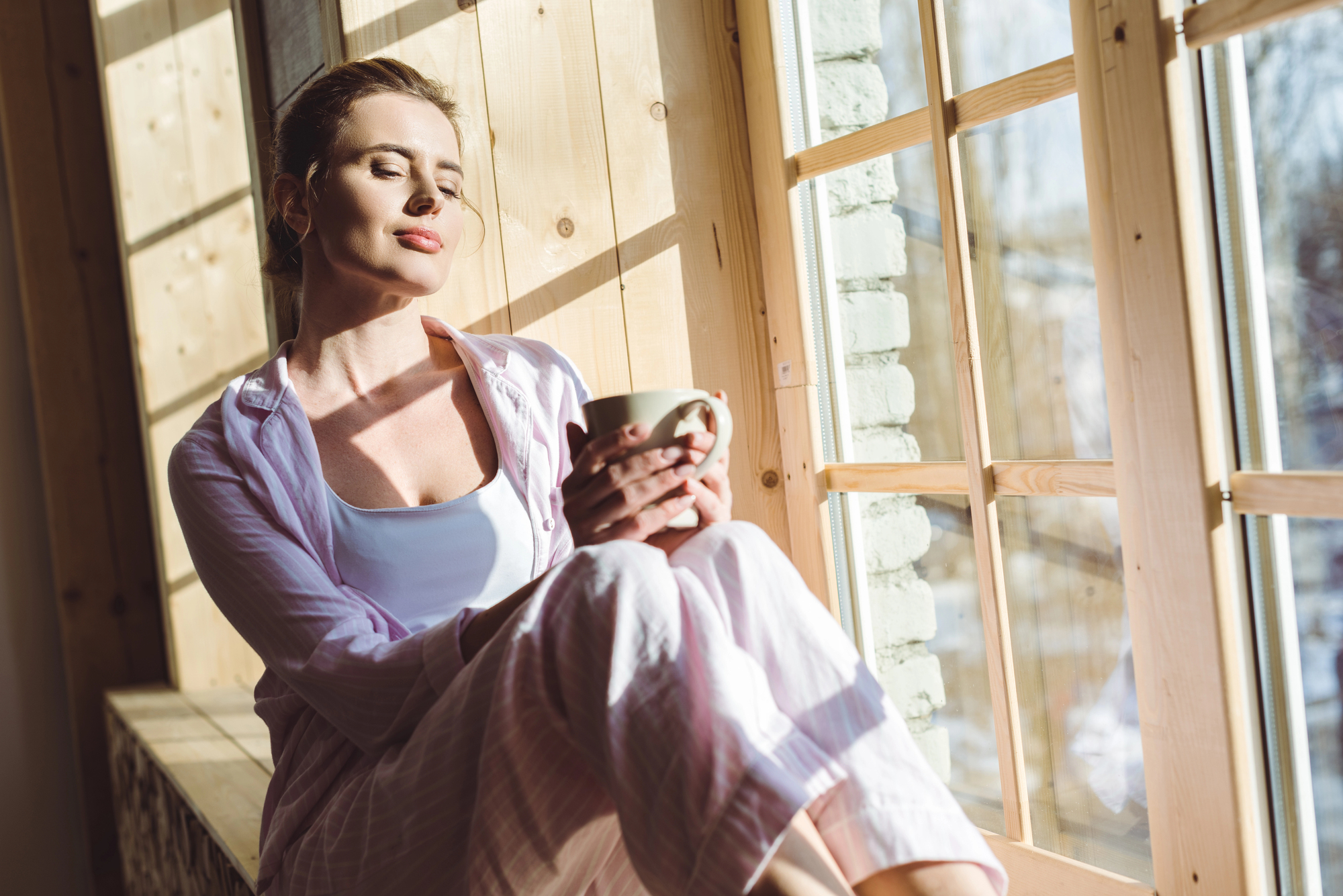 A woman in pajamas sits by a sunlit window, holding a cup of coffee or tea. She appears relaxed with her eyes closed, basking in the sunlight. The setting looks cozy with wooden walls and snow visible outside the window.