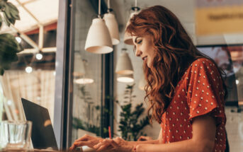 A woman with long red hair types on a laptop at a cozy café. She is wearing a red polka-dotted dress and appears focused on her work. Pendant lights hang above her, and there are plants and a glass of water nearby. The atmosphere is warm and inviting.