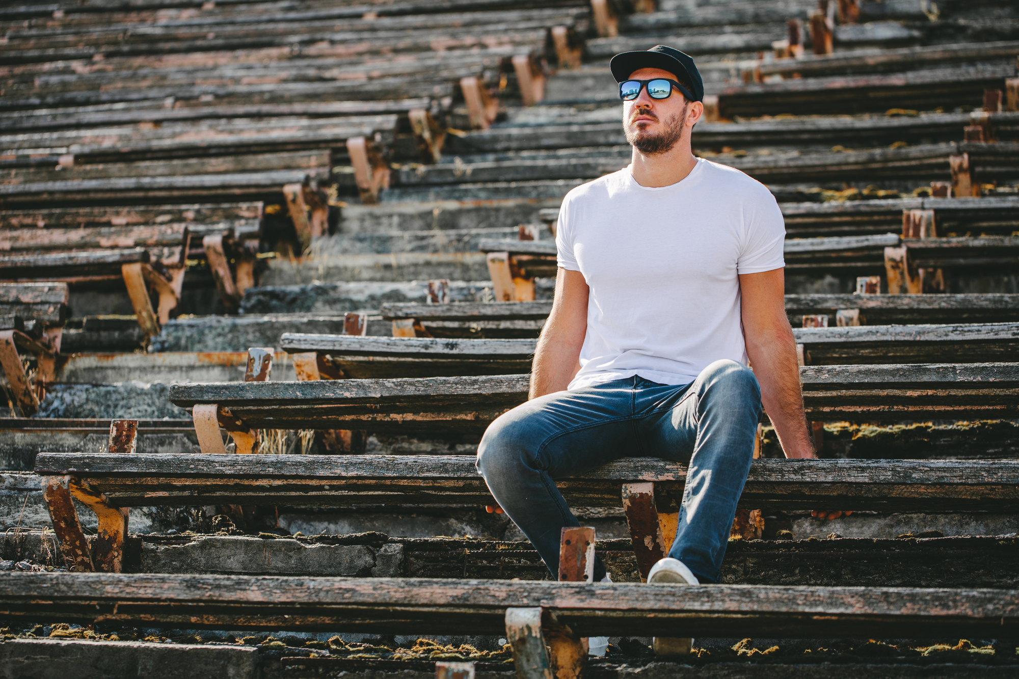 A man wearing a white t-shirt, jeans, sunglasses, and a black cap is sitting on worn wooden steps in an outdoor setting. The steps appear to be part of an old, rustic structure. The man looks up towards the sky with a contemplative expression.