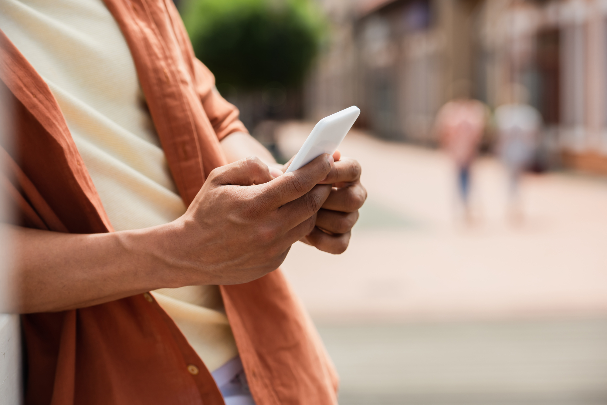 A person stands outdoors, leaning against a wall and using a smartphone. They are wearing a light beige shirt with a terracotta-colored jacket or overshirt. The background is slightly blurred, showing a street scene with greenery and people in the distance.