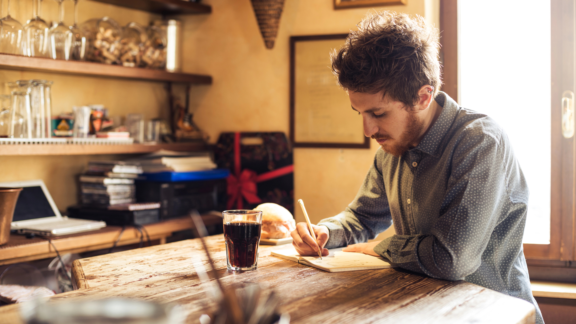 A man with brown hair and a beard sits at a wooden table by a window, writing in a notebook. He is wearing a gray shirt. A glass of a dark beverage sits on the table, along with other items like books, a bread roll, and various kitchenware in the background.