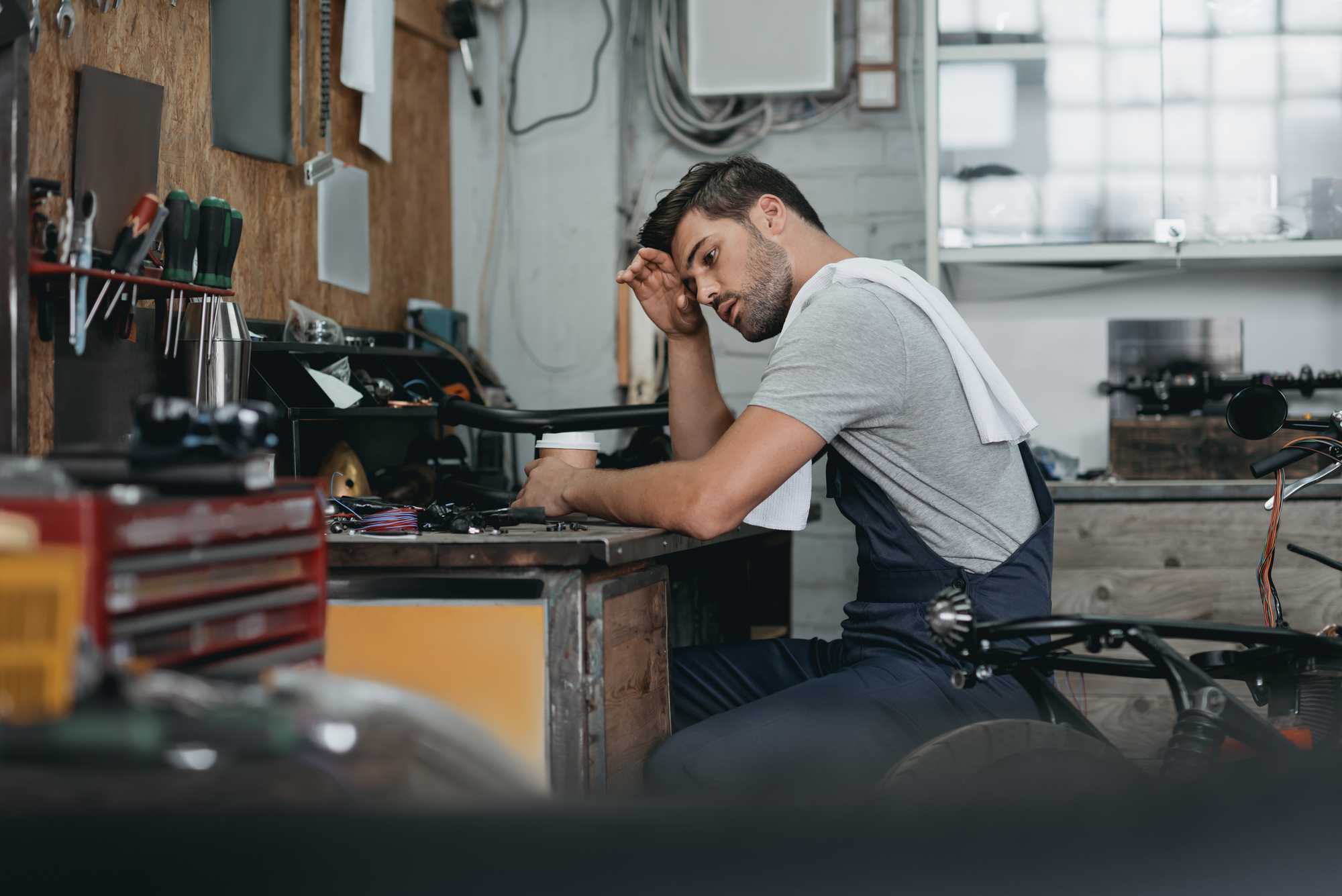 A male mechanic wearing a grey t-shirt and blue overalls sits at a cluttered workbench in a dimly lit garage. He looks tired, wiping sweat from his forehead with a white towel draped over his shoulder. Various tools and equipment are scattered around the workspace.