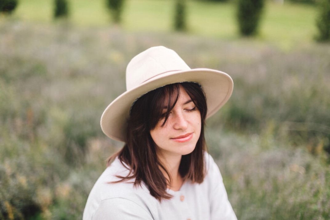 A woman with shoulder-length brown hair wearing a wide-brimmed beige hat and a light-colored shirt is sitting outdoors on a grassy field. Her eyes are closed, and she has a peaceful, relaxed expression on her face. Blurred greenery is visible in the background.