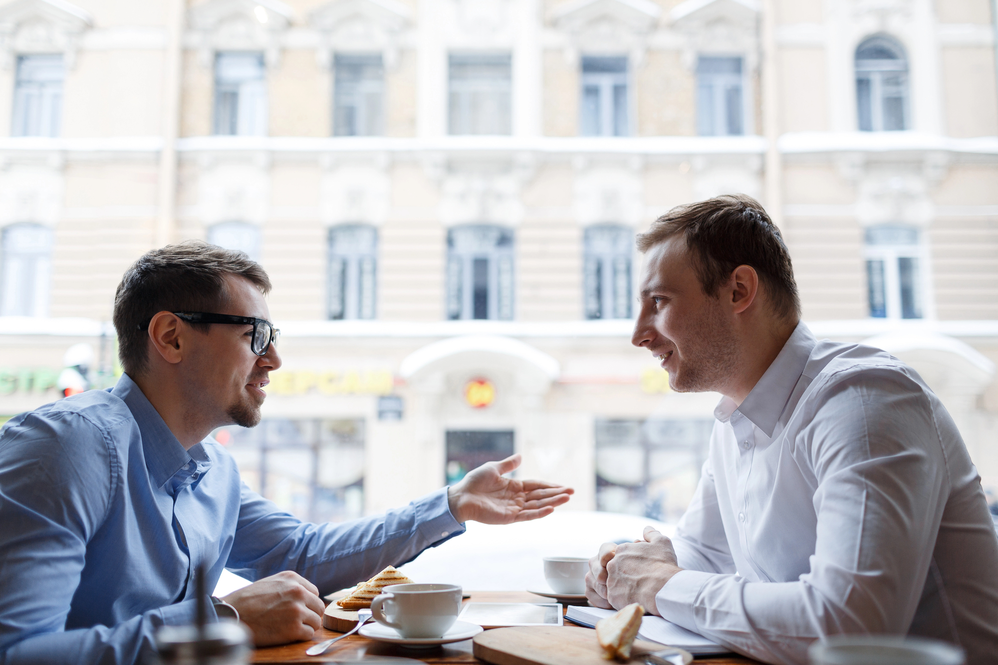 Two men sit at a table in a cafe, talking over coffee. One man, wearing glasses and a blue shirt, gestures with his hand, while the other man, in a white shirt, listens attentively. The background shows a blurred view of buildings through the large window.