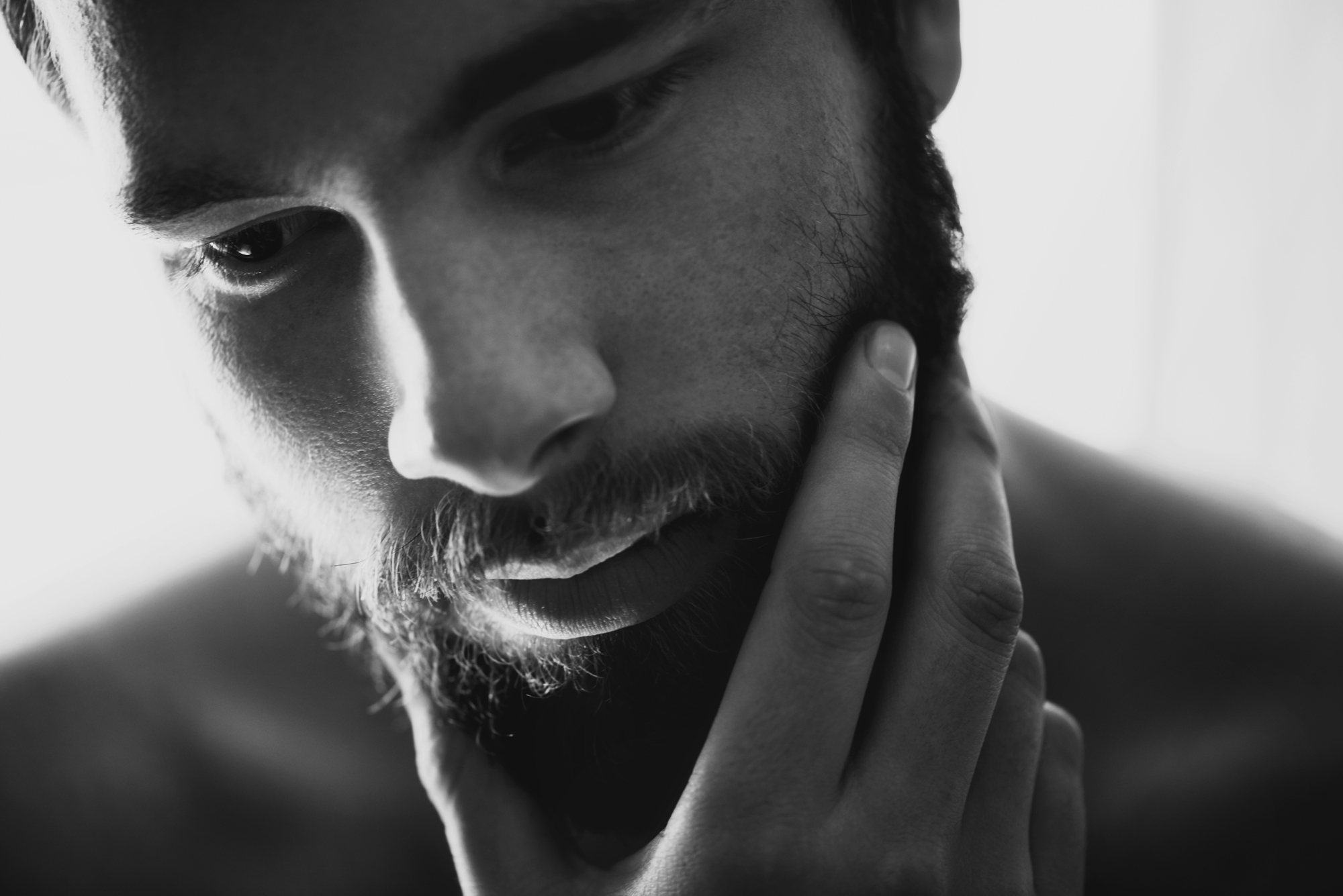 Black and white close-up of a contemplative man with a beard touching his chin. The lighting highlights his thoughtful expression, casting soft shadows on his face. Only his shoulders and upper chest are visible, with a blurred background.