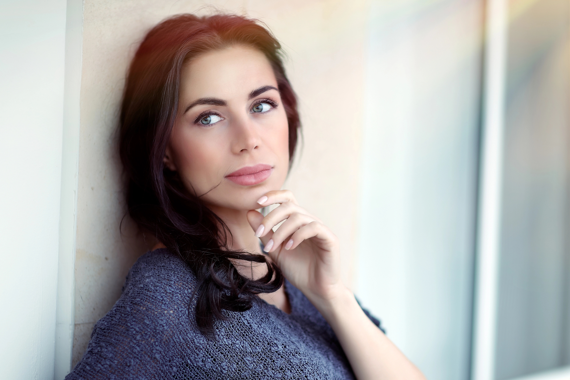 A woman with long brown hair and blue eyes is leaning against a white wall. She is wearing a gray knit sweater and has her hand gently resting on her chin, gazing thoughtfully into the distance. The background is softly lit with natural light from a nearby window.