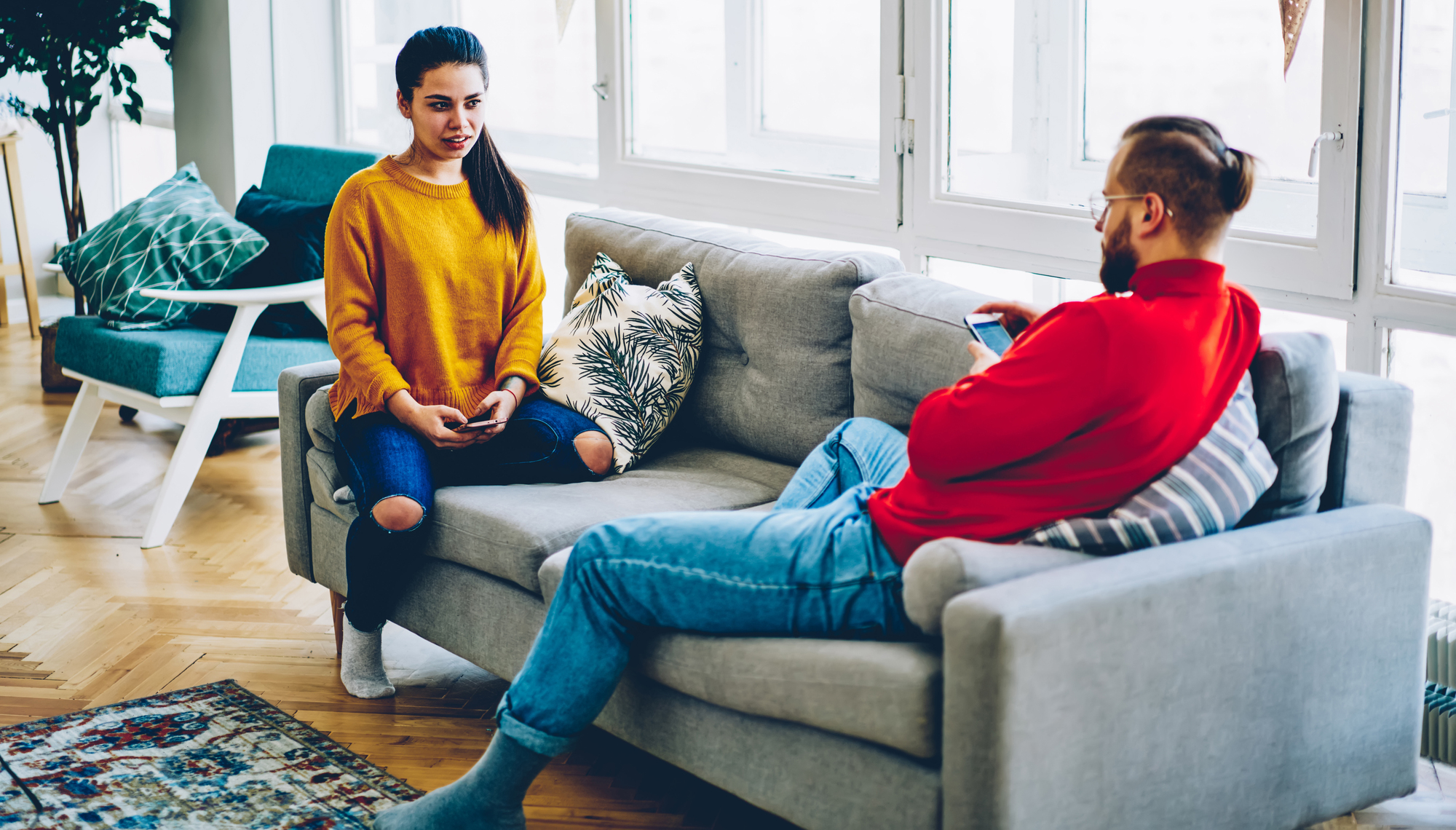 Two people are sitting in a modern living room. A woman in a yellow sweater and ripped jeans sits on the floor, leaning against a gray couch, while a man in a red sweater and jeans lounges on the couch, looking at his phone. They appear to be in a conversation.