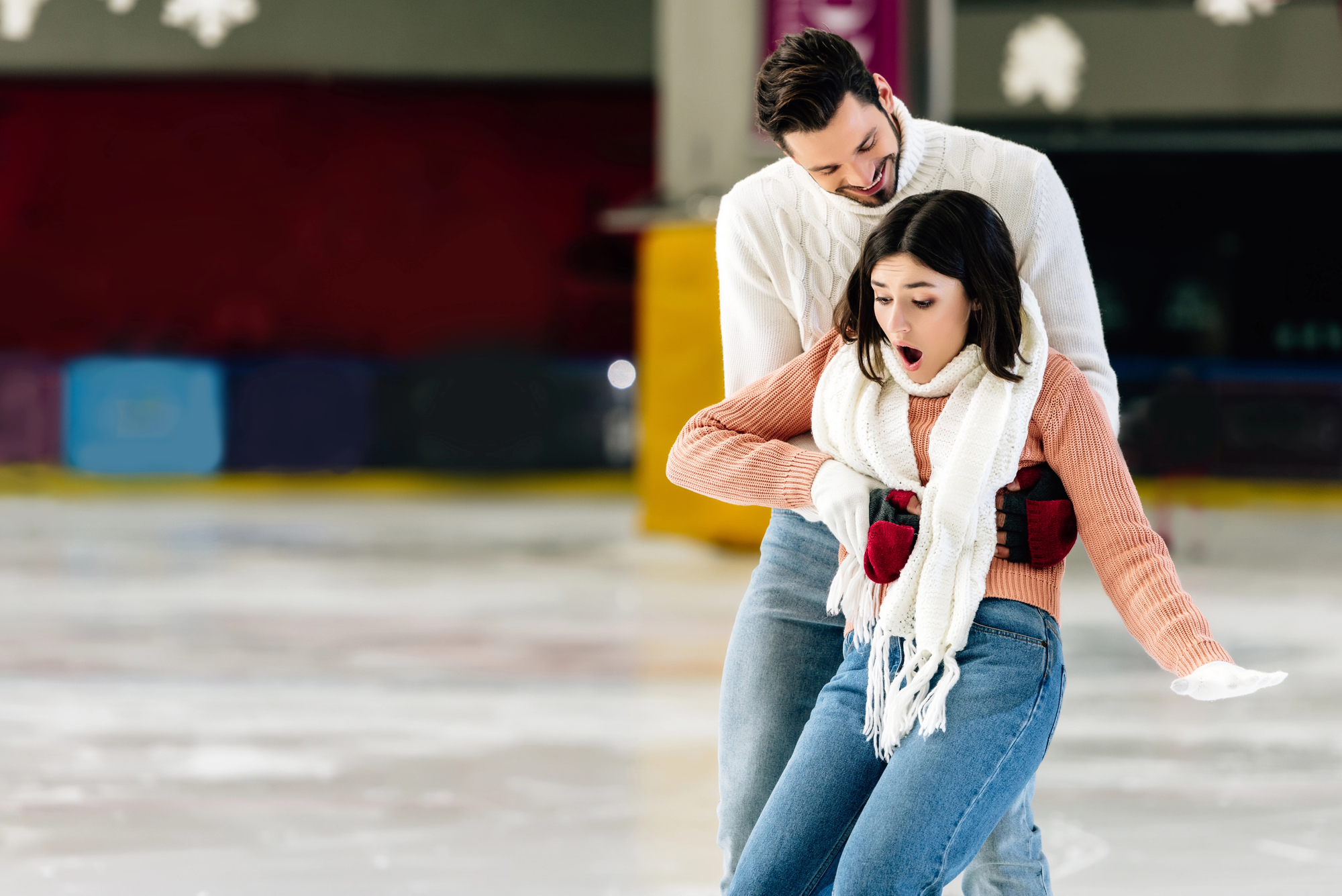 A young woman wearing a white scarf, orange sweater, and blue jeans appears surprised as she is steadied by a man in a white sweater while ice skating. They are on an indoor skating rink, and the man, who is positioned behind her, is helping her balance.