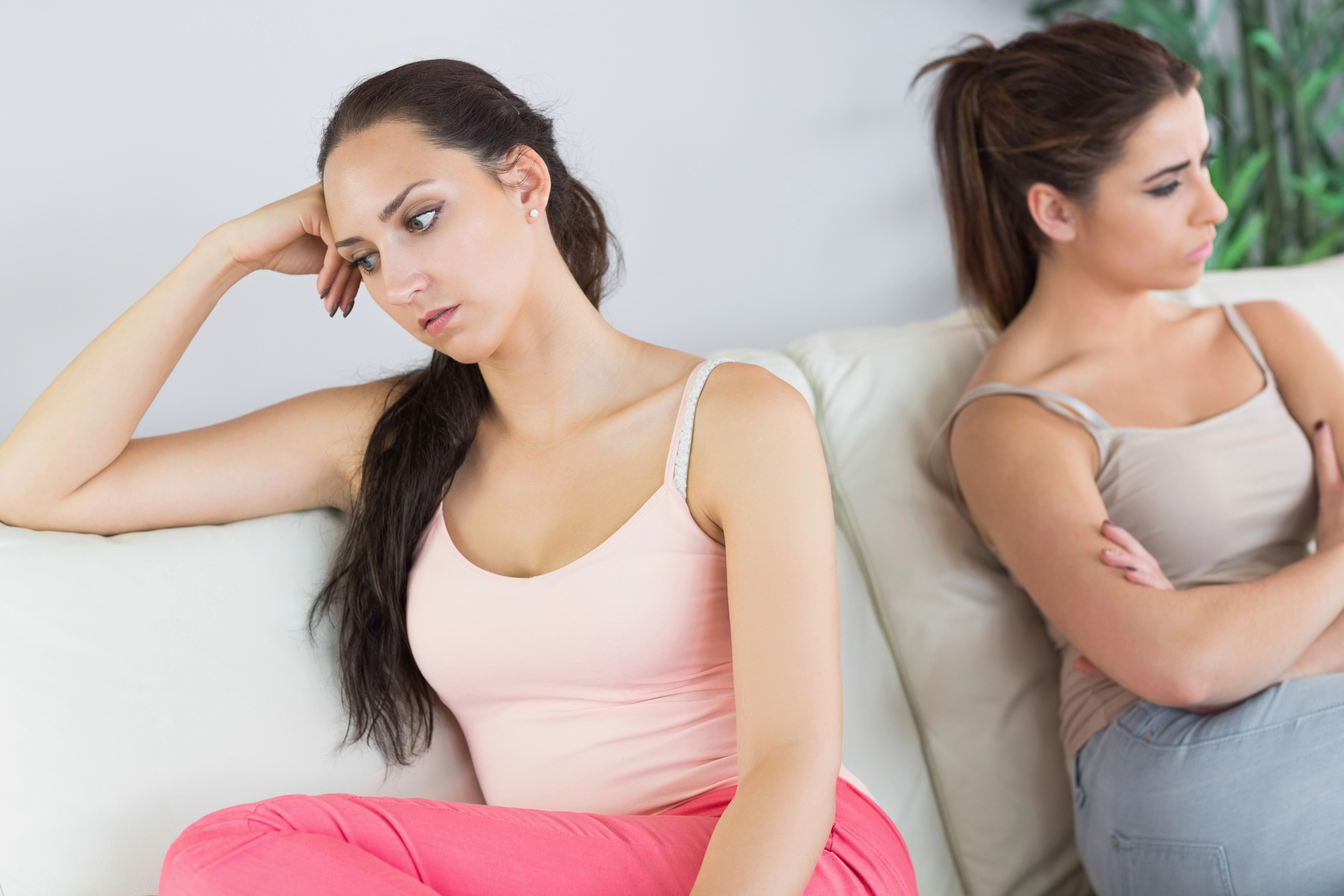 Two women sit on a white couch, both looking away from each other with upset expressions. The woman in the foreground has her head resting on her hand, wearing a pink tank top and red pants, while the other woman sits with her arms crossed, wearing a beige tank top.