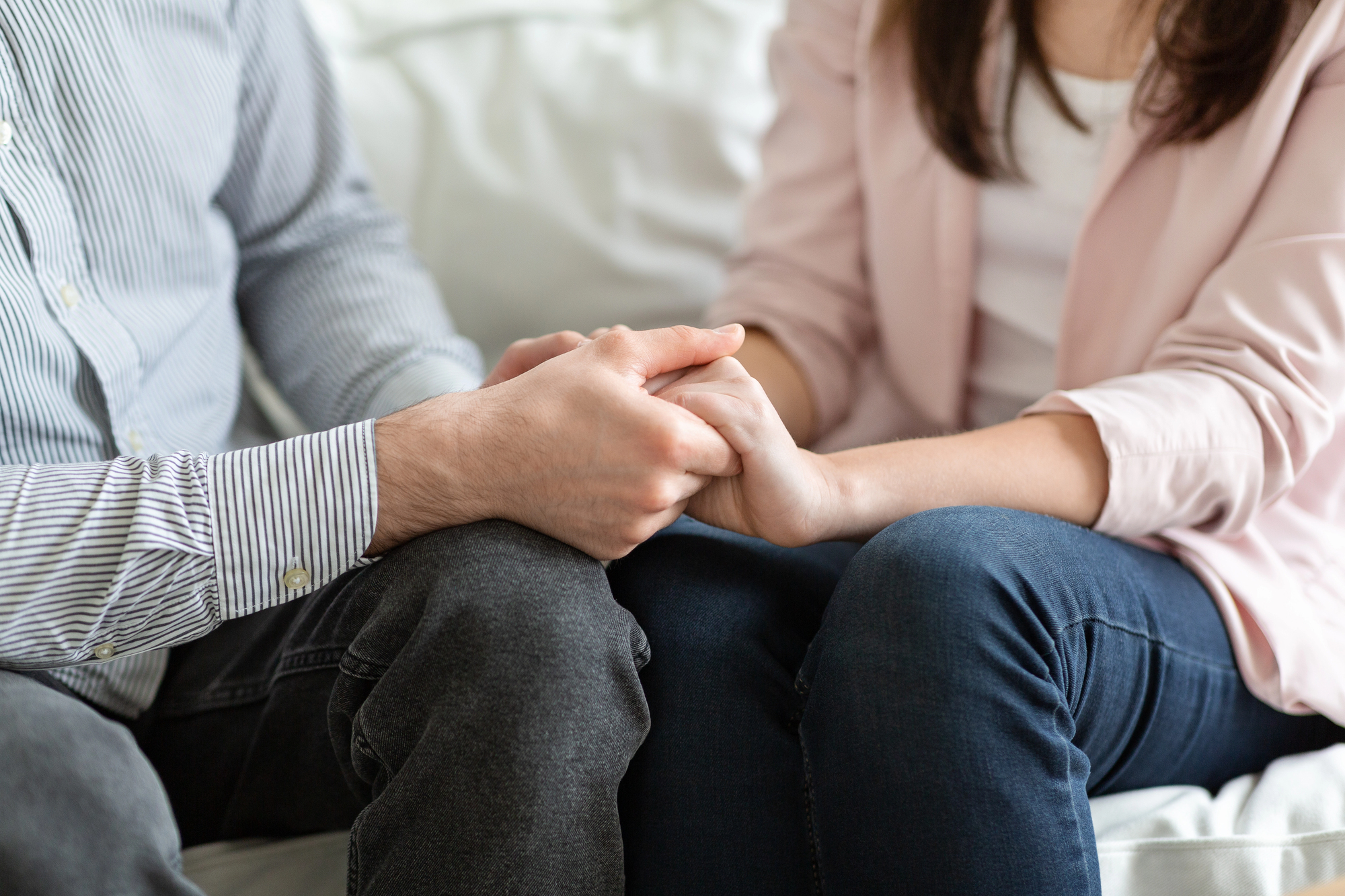 A close-up of two people sitting on a couch holding hands. One person wears a long-sleeved striped shirt and jeans, and the other wears a light jacket and jeans. They are sitting closely together, providing support and comfort to each other.