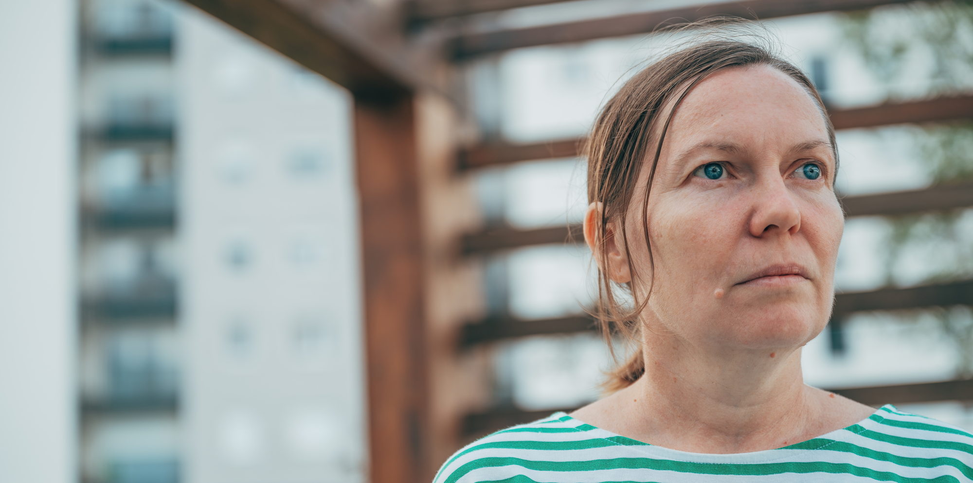 A woman with a serious expression stands outdoors. She has her hair tied back and is wearing a green and white striped shirt. In the background, there is a partially blurred view of buildings and a structure made of wooden beams.