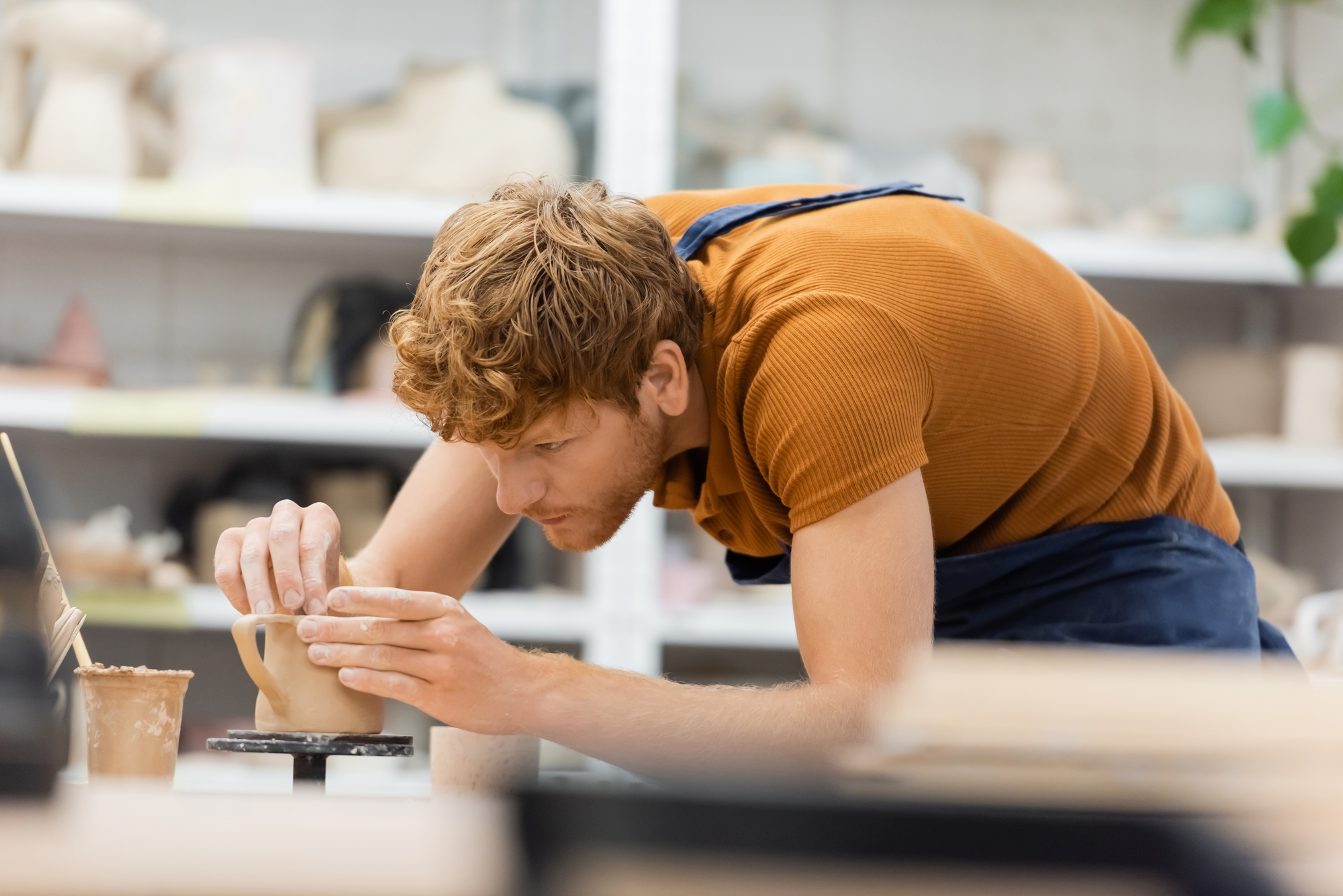 A person with curly red hair, wearing an orange shirt and dark apron, is focused on shaping a clay pot on a pottery wheel. Shelves of pottery and art supplies are visible in the background.