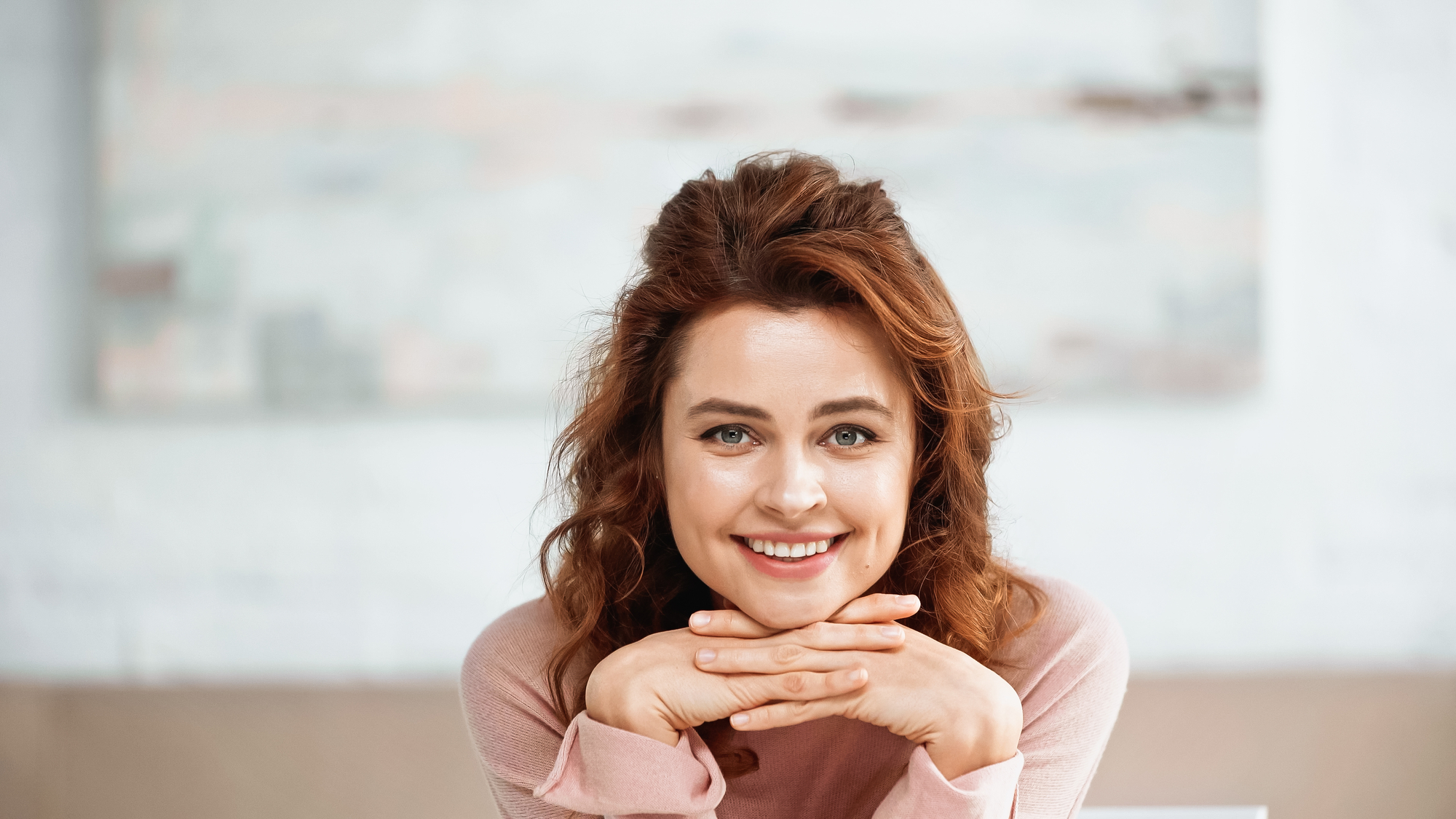 A woman with reddish-brown hair smiles at the camera, resting her chin on her hands. She is wearing a light pink sweater and is positioned in front of a blurred, light-colored background.