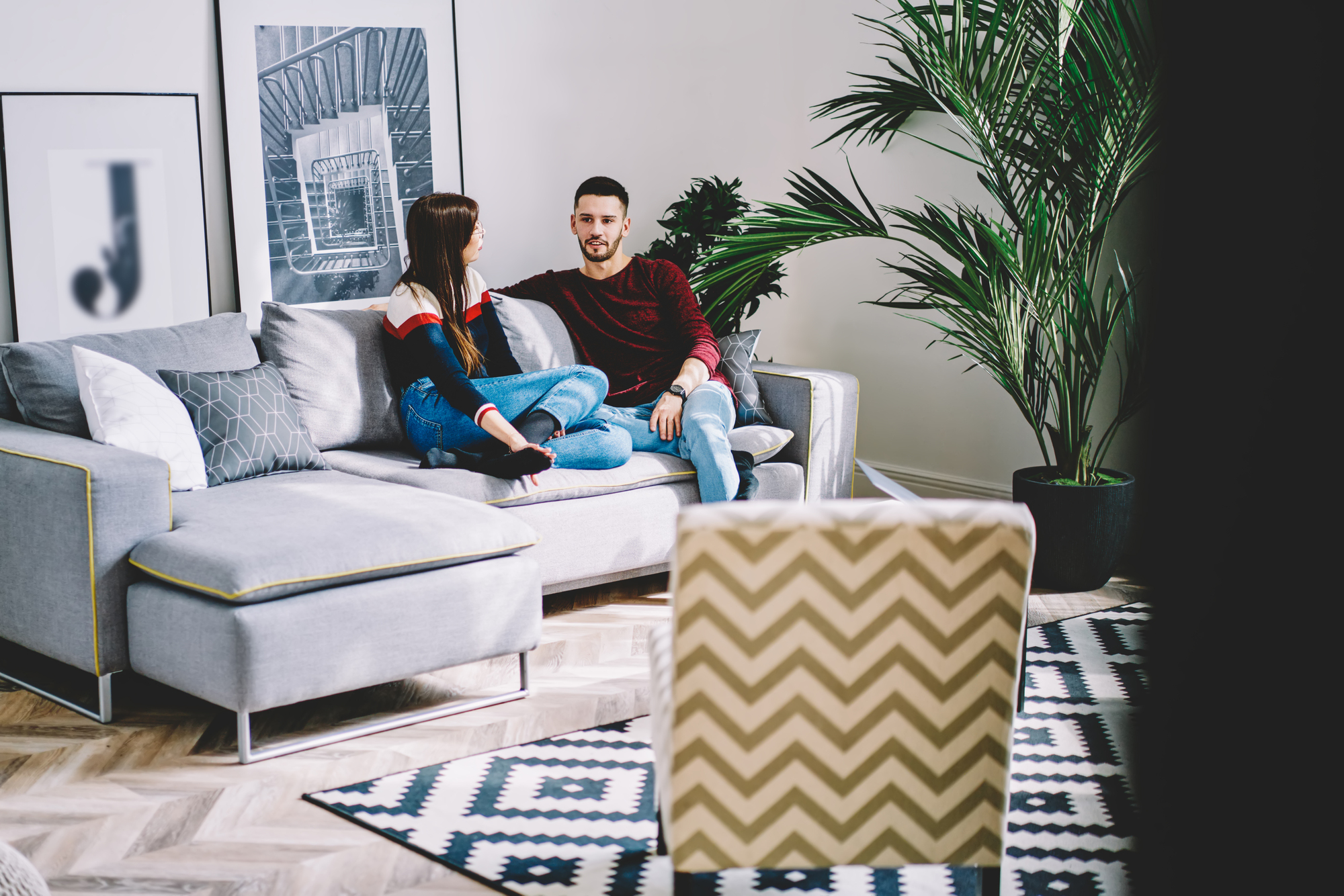 A man and woman sit together on a modern grey sofa in a stylish living room. The room features a geometric rug, large green plant, framed wall art, and scattered cushions. The couple appears relaxed and engaged in conversation.