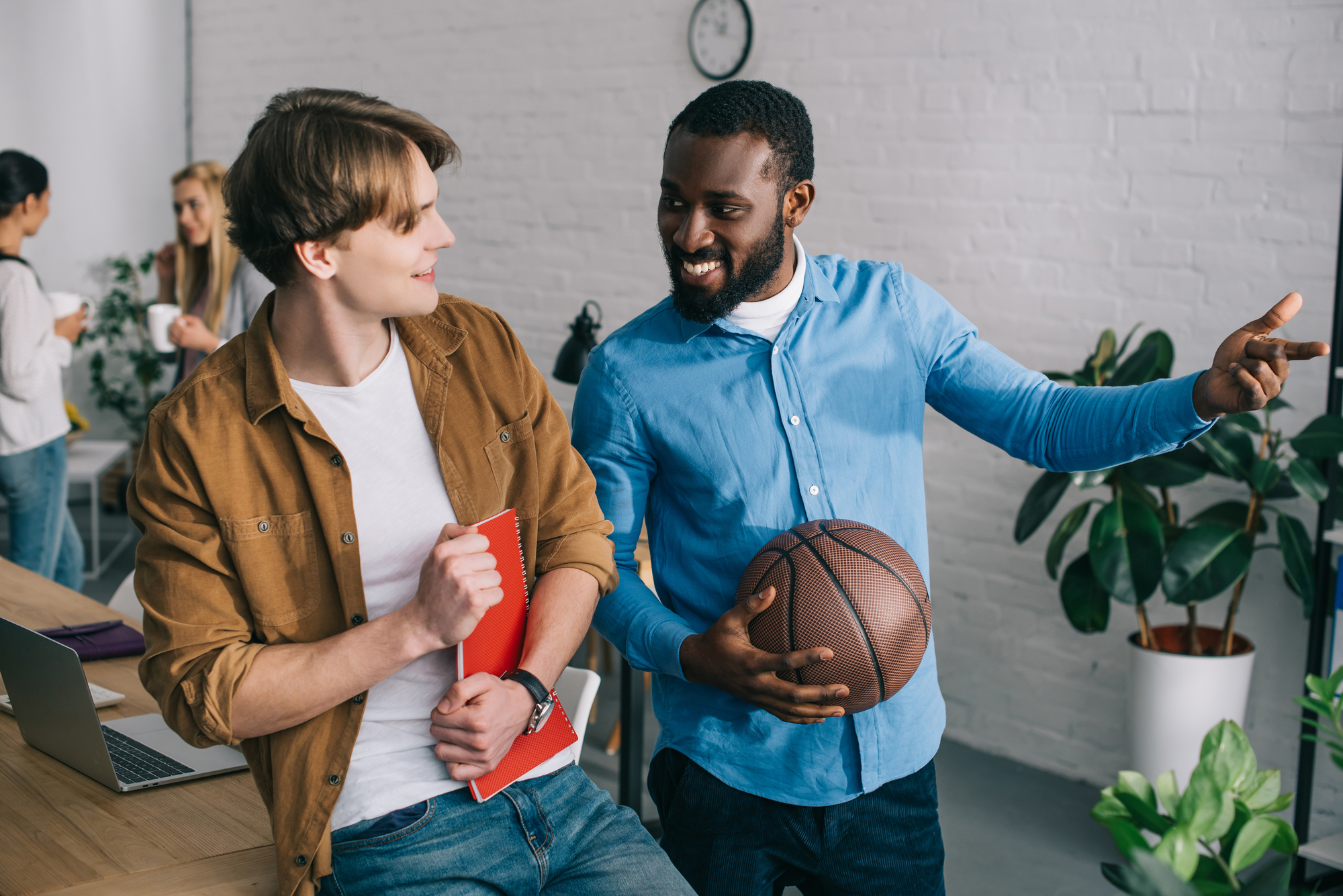 Two men are casually chatting in an office. The man on the left is holding a red notebook, while the man on the right is holding a basketball and pointing. Other people in the background appear to be engaged in conversation. The setting is relaxed and informal.