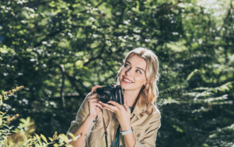 A smiling woman with blond hair stands in a lush, green forest. She is holding a camera, poised to take a photo. She is wearing a khaki jacket and appears to be enjoying the natural surroundings. Sunlight filters through the trees.