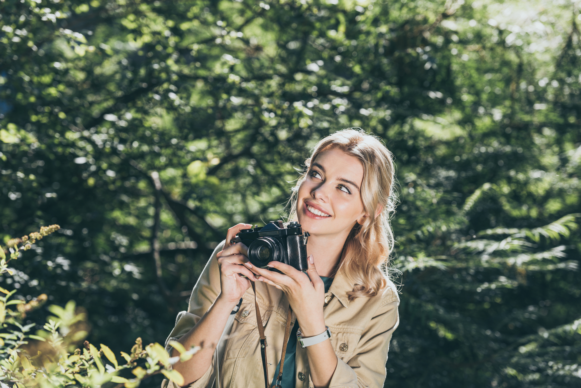 A smiling woman with blond hair stands in a lush, green forest. She is holding a camera, poised to take a photo. She is wearing a khaki jacket and appears to be enjoying the natural surroundings. Sunlight filters through the trees.