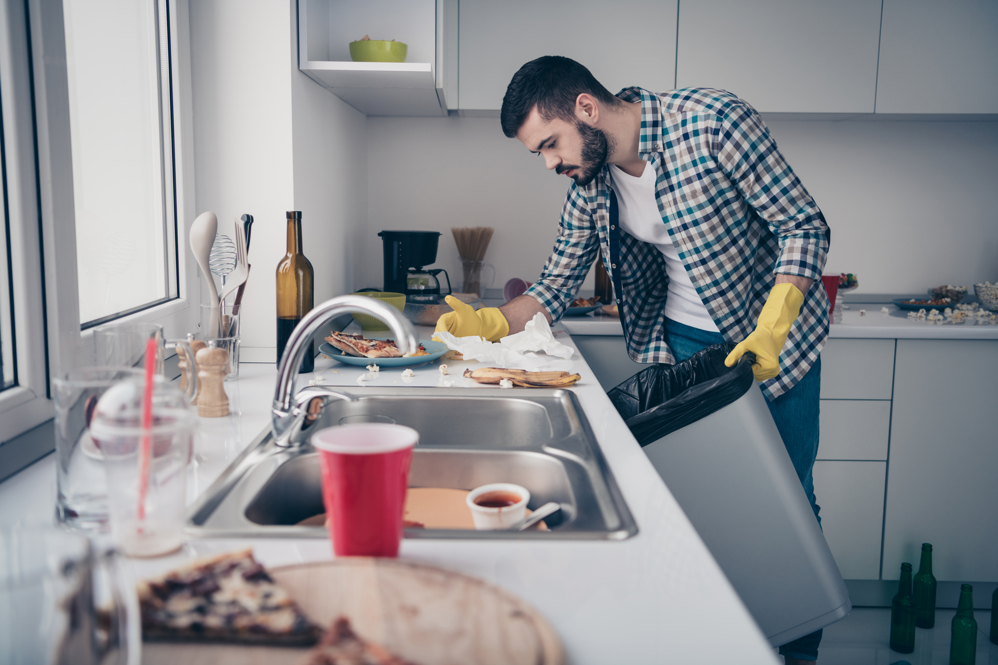 A bearded man wearing yellow rubber gloves is cleaning a modern white kitchen. He is leaning over a black trash bin, throwing away pizza leftovers. The countertop is cluttered with various items, including a red cup, a plate with pizza, and a coffee mug.