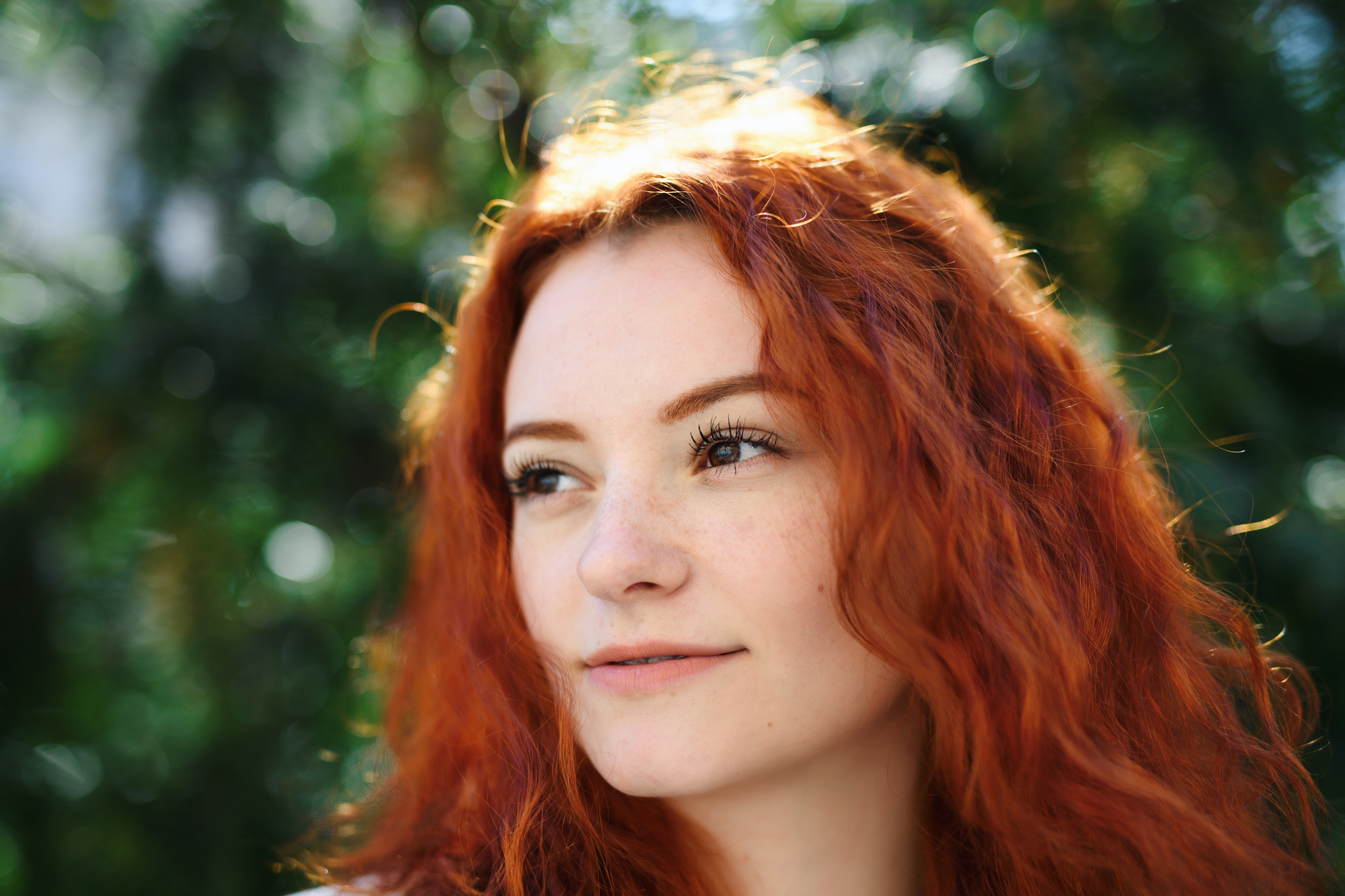 A person with long, wavy red hair is shown in a close-up, outdoor portrait. They are gazing slightly to the right with a soft smile. The background is blurred, featuring green foliage and sunlight filtering through.