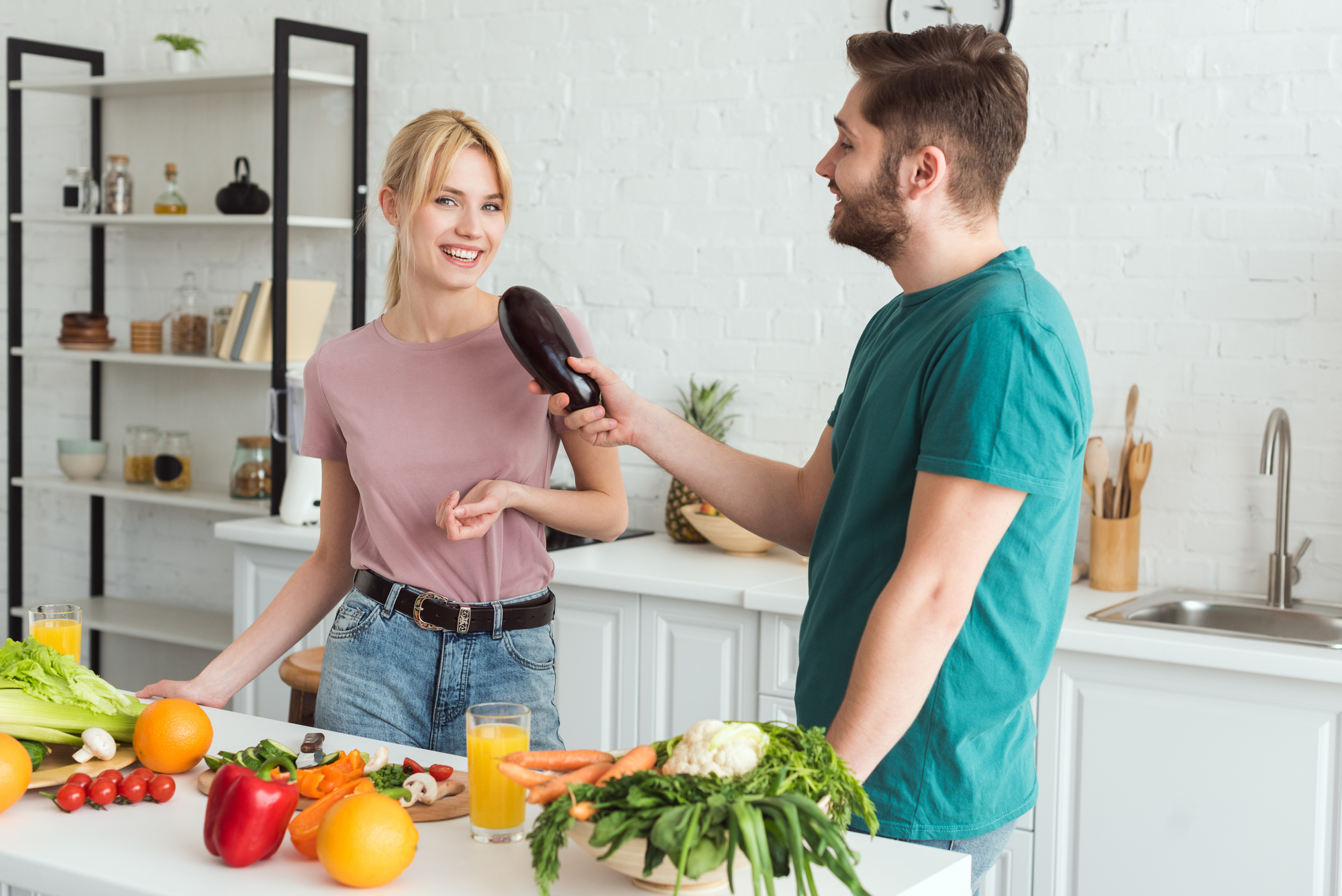 A woman and a man are in a bright, modern kitchen preparing a meal. The woman, smiling, hands on the counter, looks at the man who holds an eggplant towards her. Fresh vegetables and glasses of orange juice are on the counter. Shelves with jars are in the background.