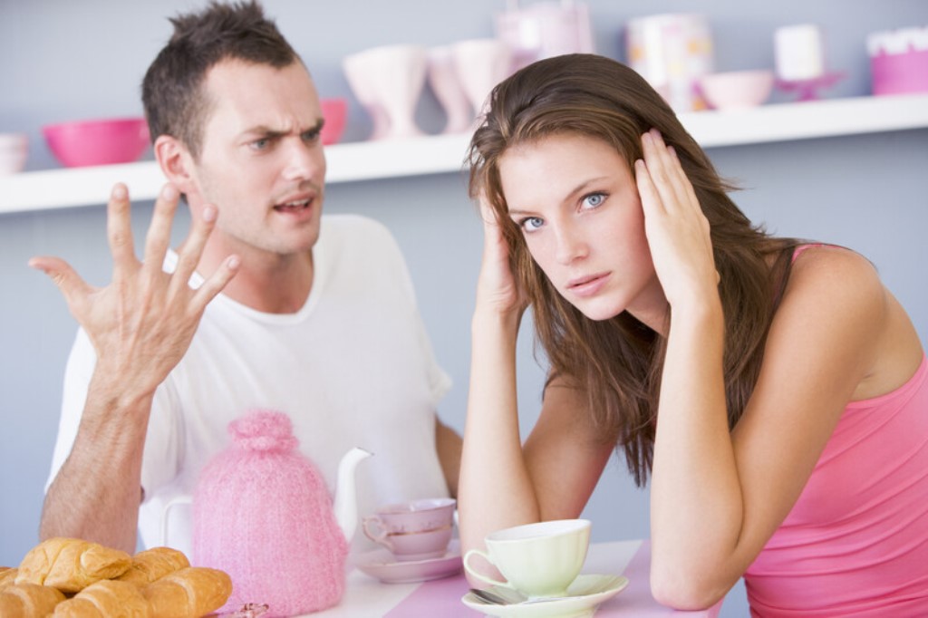 A woman sits at a table with a displeased expression, holding her head with both hands. A man beside her is gesturing with one hand, appearing to be upset or arguing. On the table are a teapot with a pink cozy, cups, and croissants. A shelf with items is seen in the background.