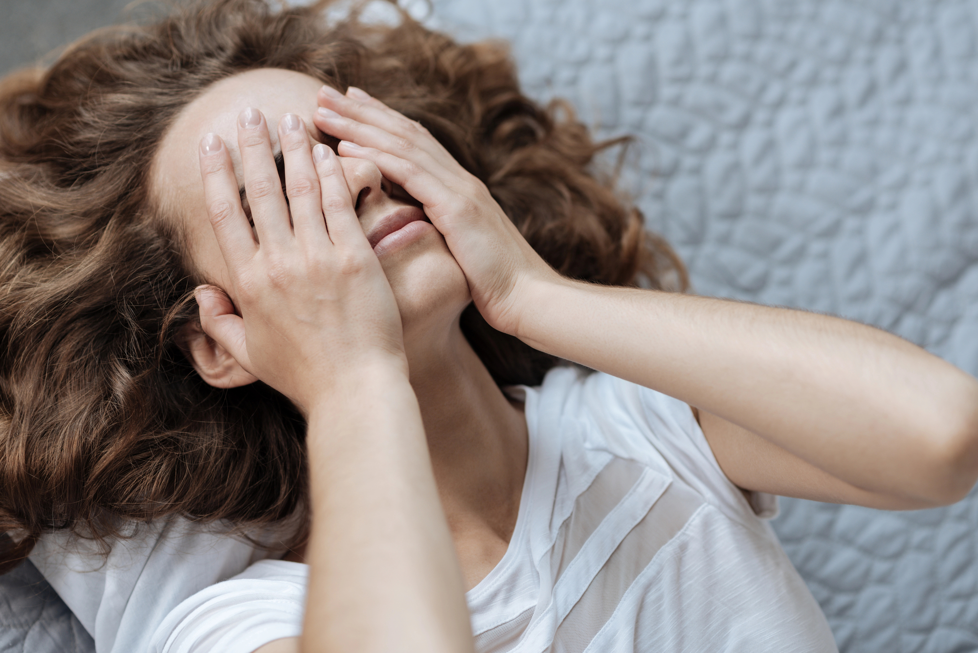 A person with long, curly hair is lying down with both hands covering their face. They are wearing a white t-shirt, and the background shows a textured, light-colored surface. The person's expression suggests they might be tired or stressed.