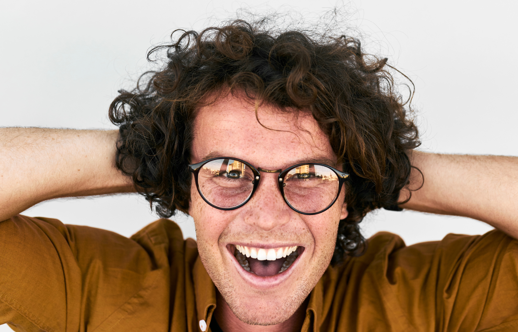 A person with curly hair and glasses smiles broadly with an open mouth. They are holding their hands behind their head and wearing a brown shirt. The background is plain and light-colored.