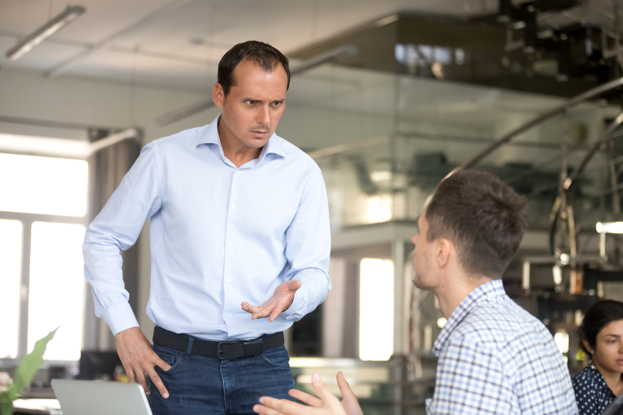 A man in a light blue shirt stands with a serious expression, gesturing with one hand as he speaks to another man seated in front of him wearing a checkered shirt. They are in an office environment with glass partitions and a woman seated in the background.