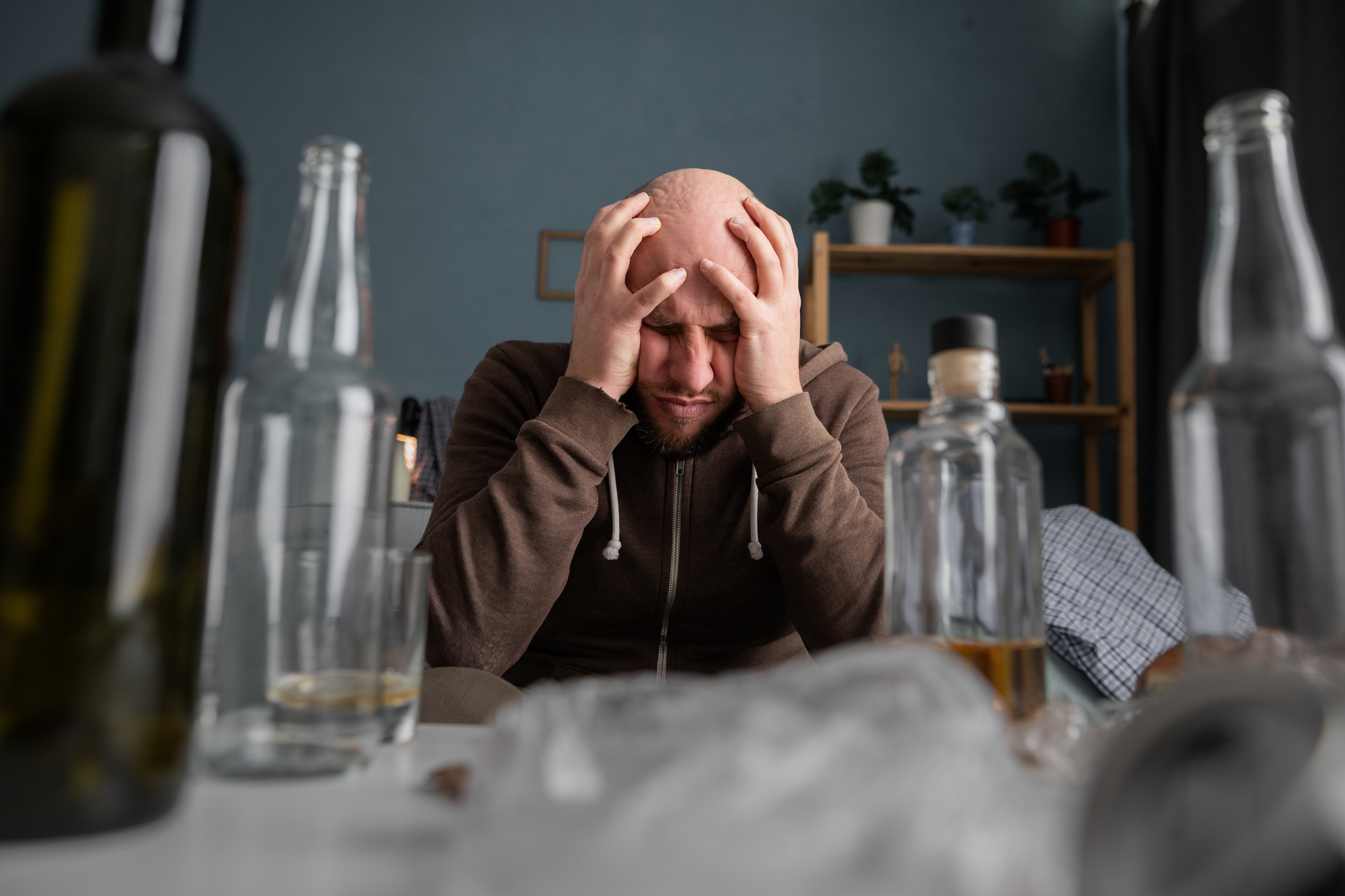 A man with a bald head and beard sits at a table, clutching his head in frustration or despair. Around him are several empty, half-empty, and full bottles, suggesting a setting of distress or overconsumption. The background includes household items and plants.