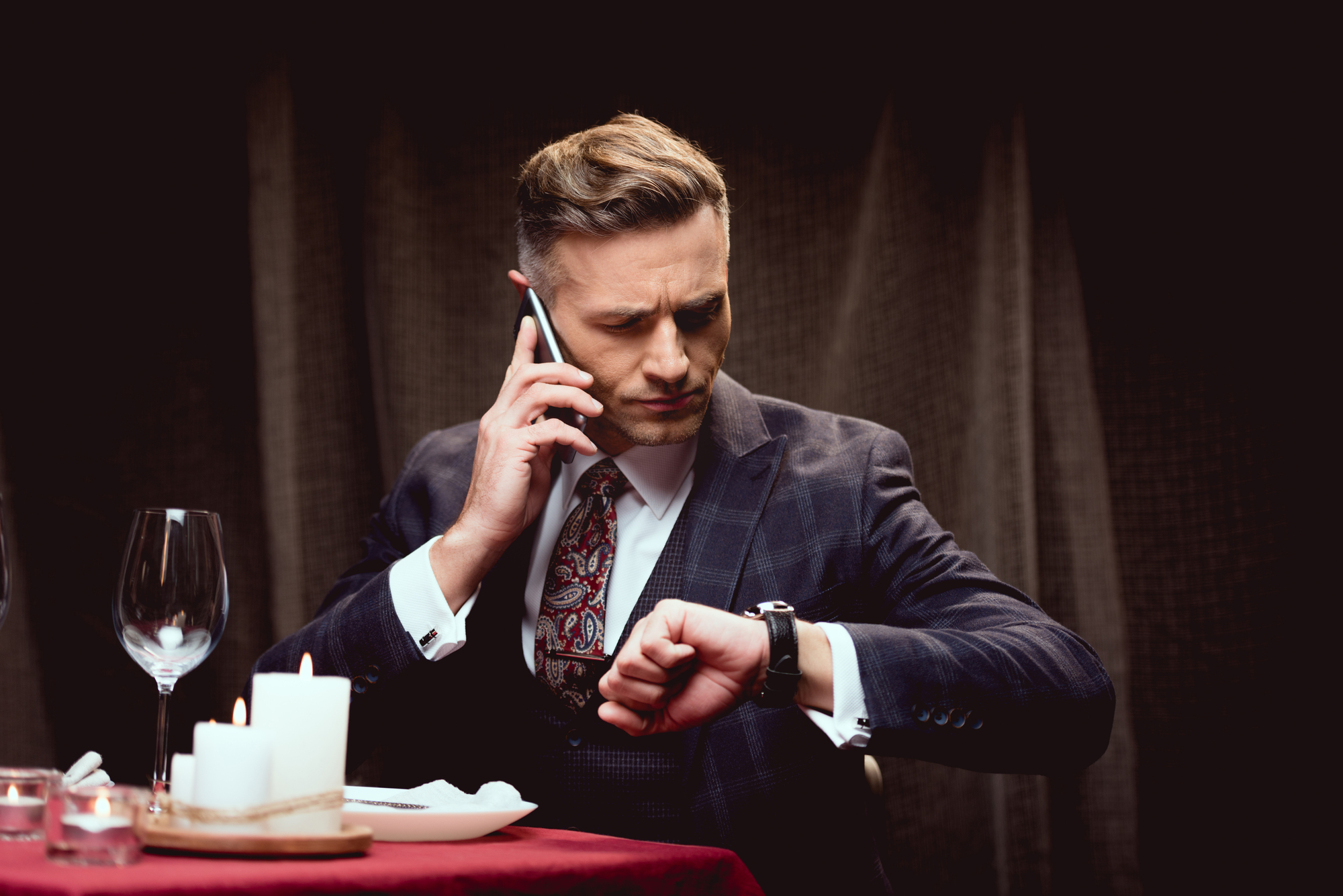 A man in a suit sits at a dining table with red tablecloth, white candles, an empty wine glass, and plate. He is talking on a cellphone and looking at his wristwatch, appearing serious and focused.