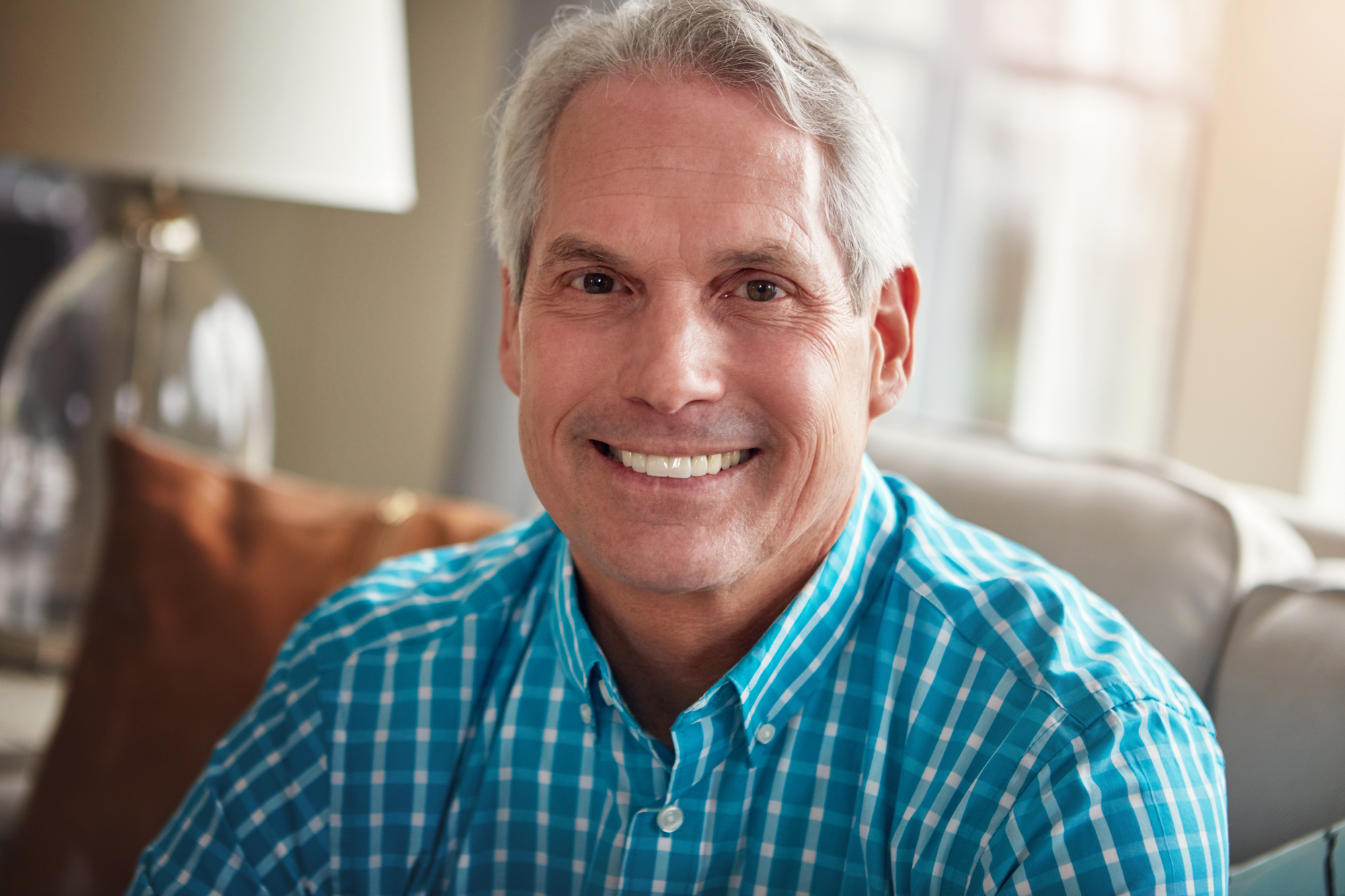 A smiling older man with short gray hair wears a blue and white checkered shirt. He is sitting indoors on a light-colored couch with a brown cushion and a table lamp in the background. The lighting is warm and natural.