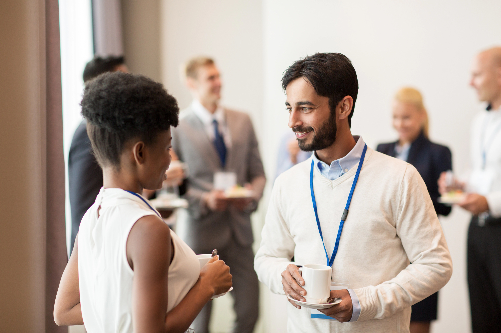 A man and a woman are conversing while holding cups of coffee, wearing blue lanyards, in a professional setting. Other people in business attire can be seen in the background engaging in discussions and holding beverages.