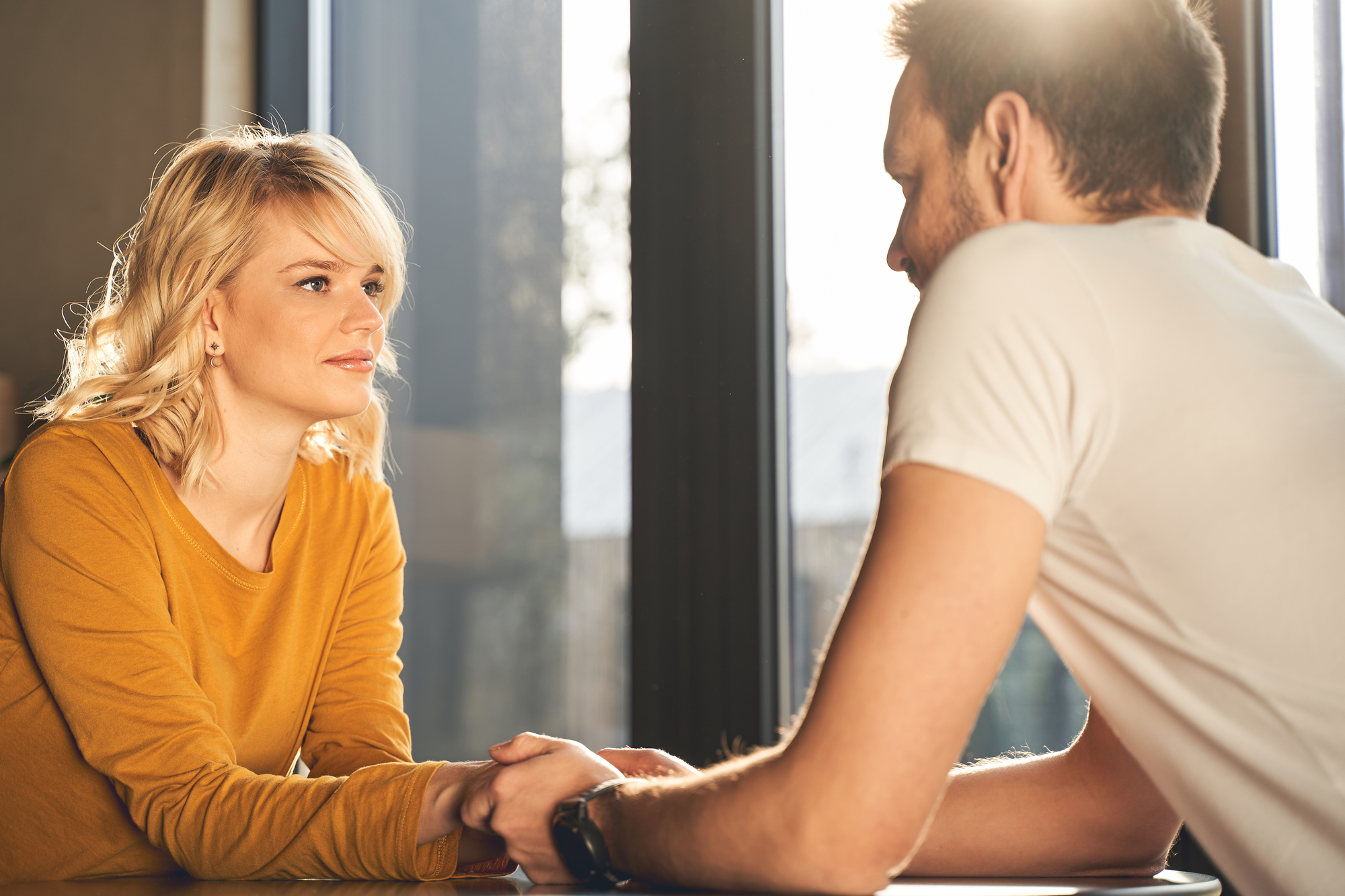 A woman with blonde hair wearing a mustard-colored shirt and a man with short hair and a white T-shirt sit across from each other at a table. They hold hands and gaze at each other, with sunlight streaming in from the window behind them.