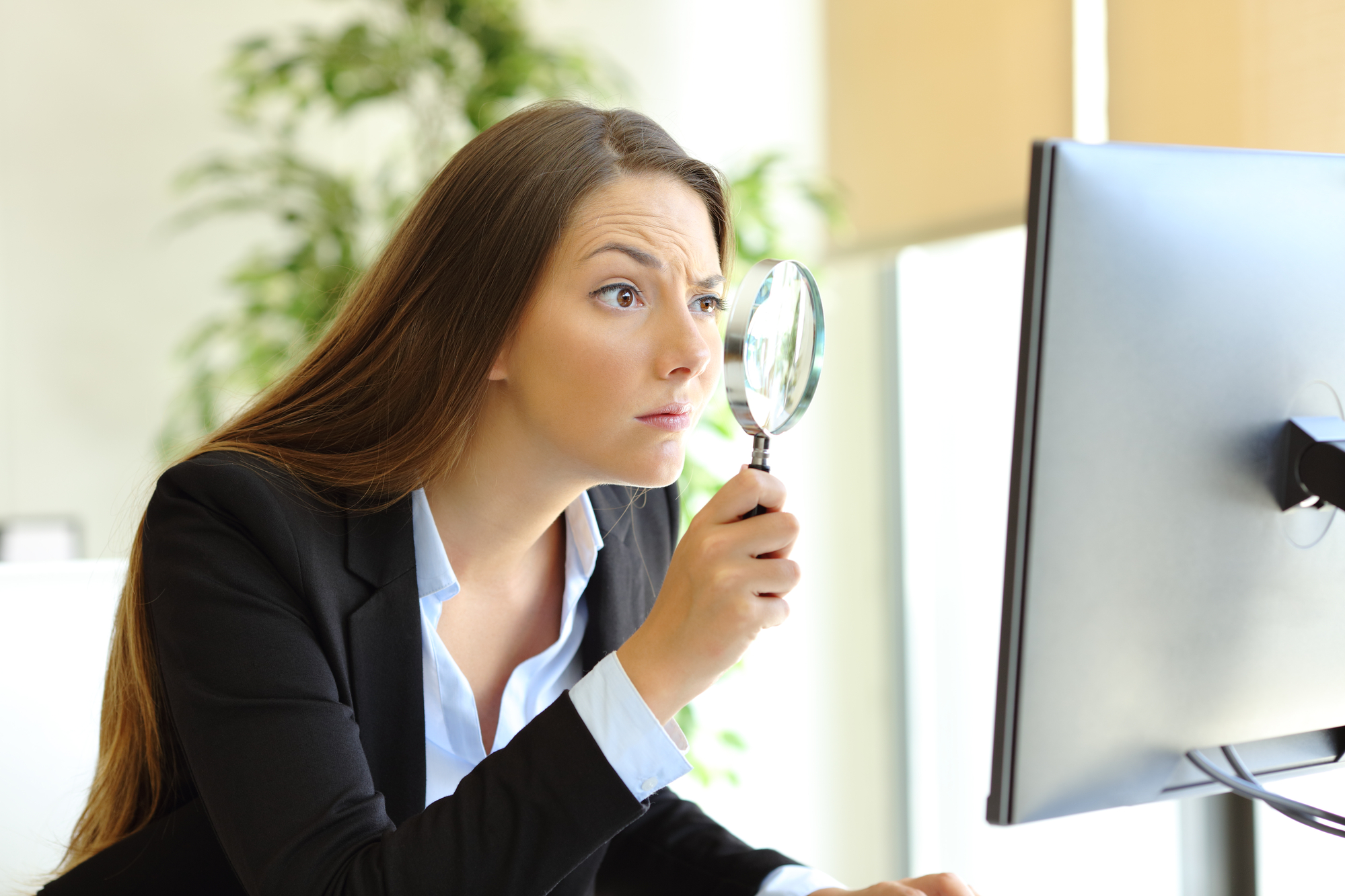 A woman in a business suit is sitting at a desk, holding a magnifying glass up to a computer screen. She appears to be inspecting the screen closely. There is a large plant in the background.