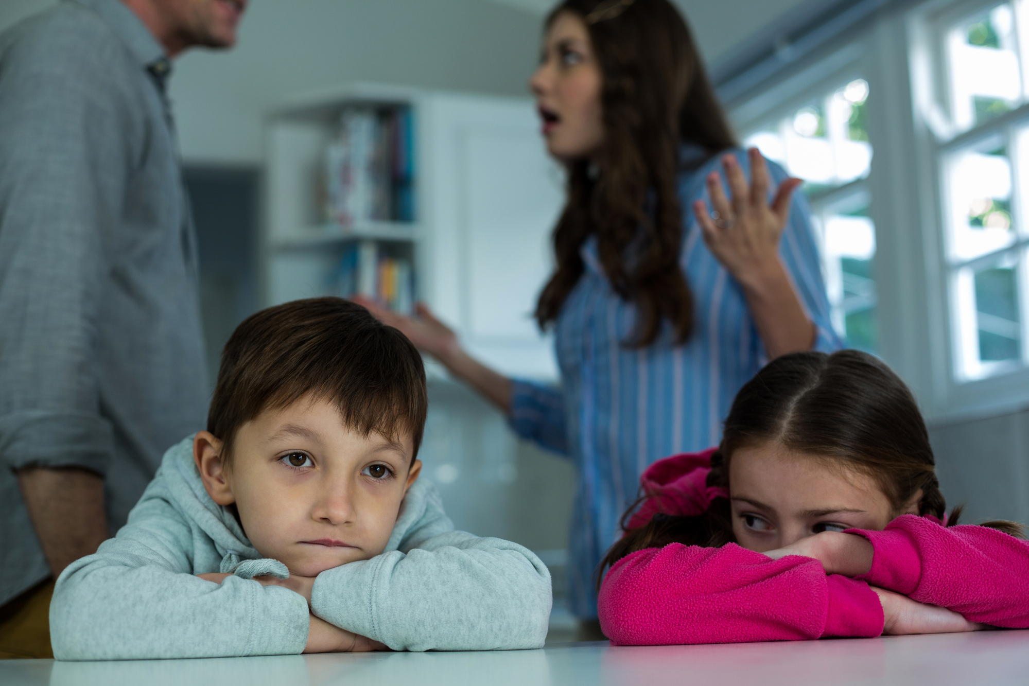 Two young children lean on a table with sad expressions while a man and woman are seen in the background arguing. The scene takes place in a brightly lit room with windows and a bookshelf. The mood is tense, reflecting family conflict.