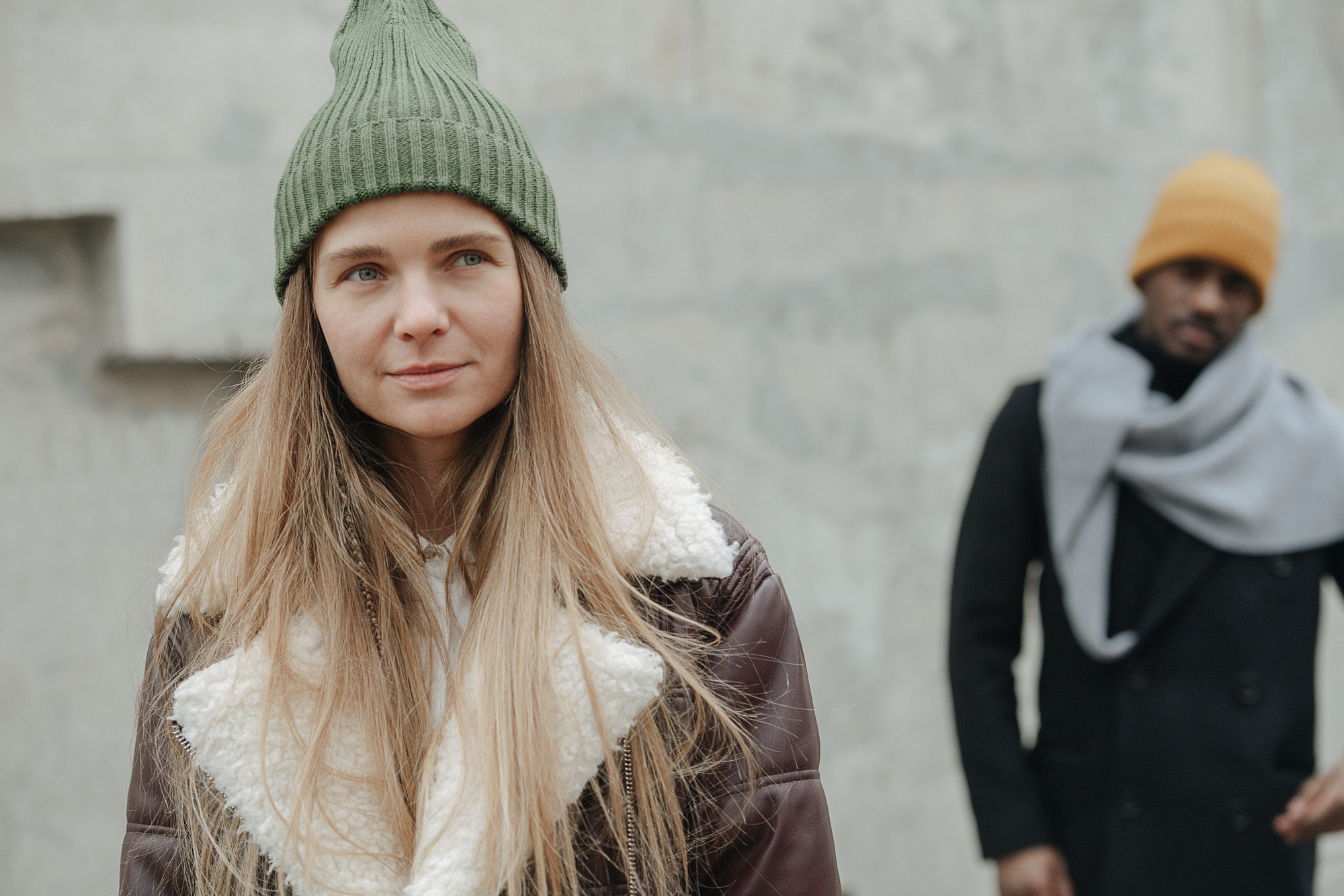 A woman with long blonde hair, wearing a green knit hat and a brown jacket with a fur collar, stands in the foreground. In the blurred background, a man wearing a yellow beanie and a grey scarf around his neck looks on. They appear to be outdoors on a chilly day.