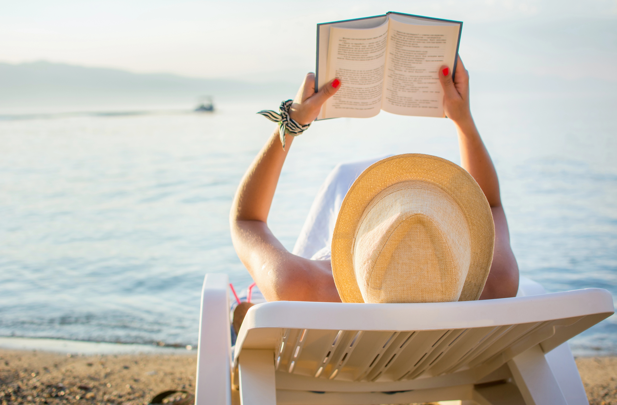 A person relaxes on a lounge chair at the beach, reading a book. They are wearing a wide-brimmed straw hat and a patterned scarf on their wrist. The ocean is calm in the background, with a faint outline of mountains and a small boat on the horizon.