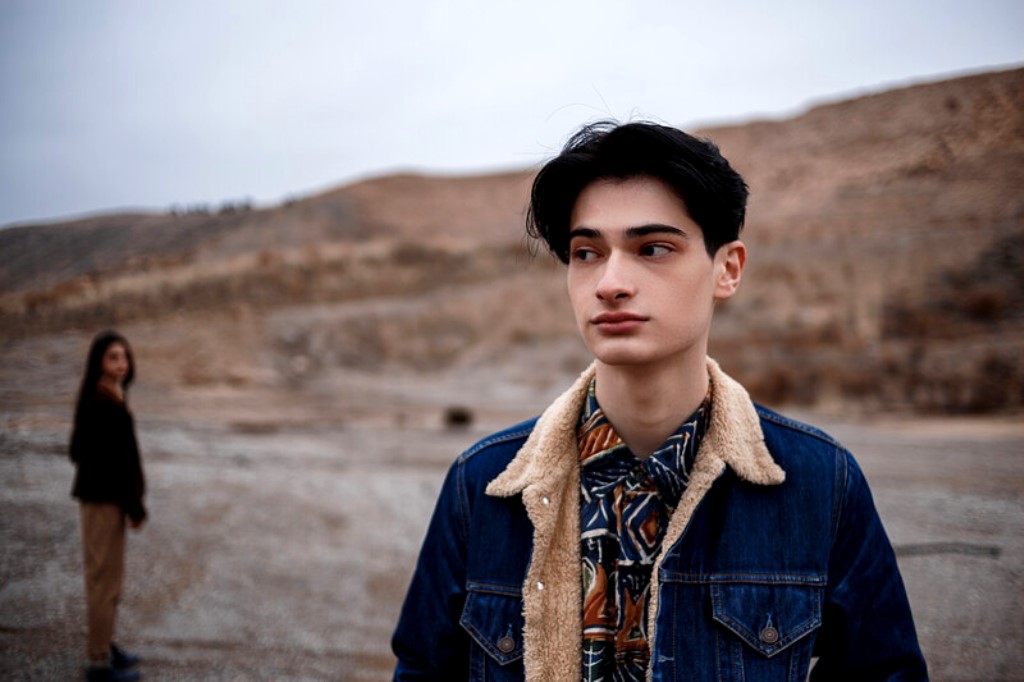 A young person with short, dark hair and dressed in a denim jacket over a patterned shirt stands in the foreground. In the blurred background, another person wearing dark clothing is partially visible in a rocky, barren landscape.