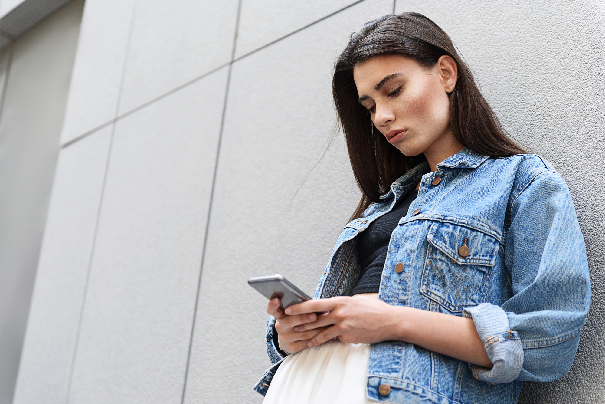 A young woman with long dark hair stands against a light gray wall, looking down at her phone. She is wearing a blue denim jacket over a black top and a light-colored skirt. She appears focused on the device in her hands.