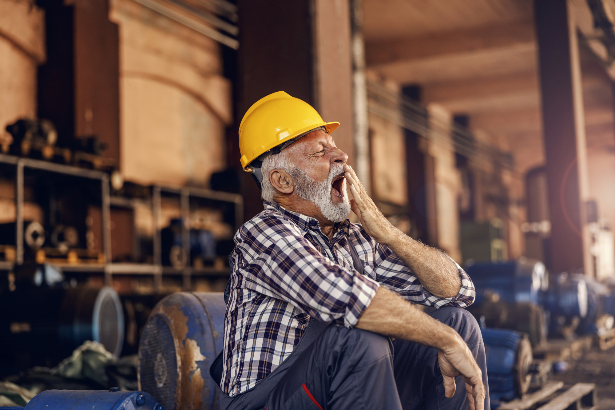 An older man wearing a yellow hard hat and plaid shirt sits in an industrial setting, yawning with his hand covering his mouth. Various machinery and equipment are visible in the background.